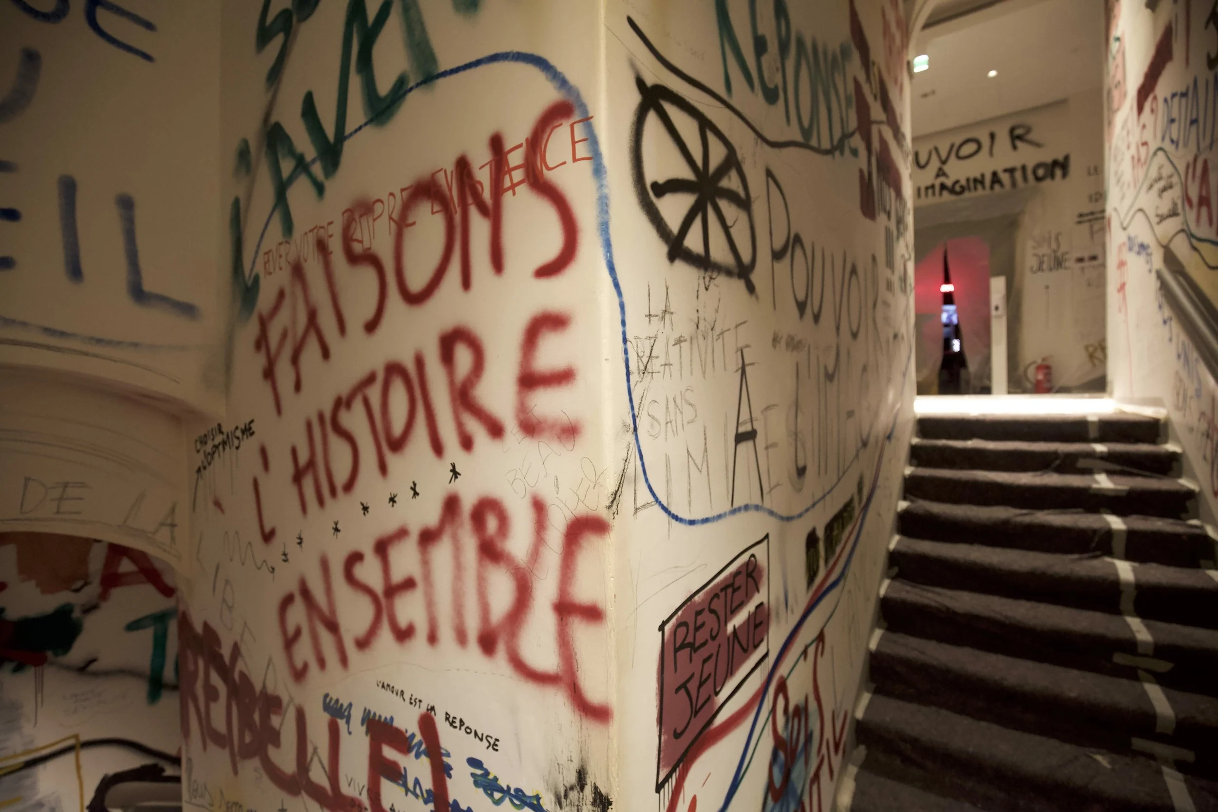 Muro decorato con graffiti e scritte in varie lingue, vicino a una scala con gradini di pietra, in un'ambientazione interna.