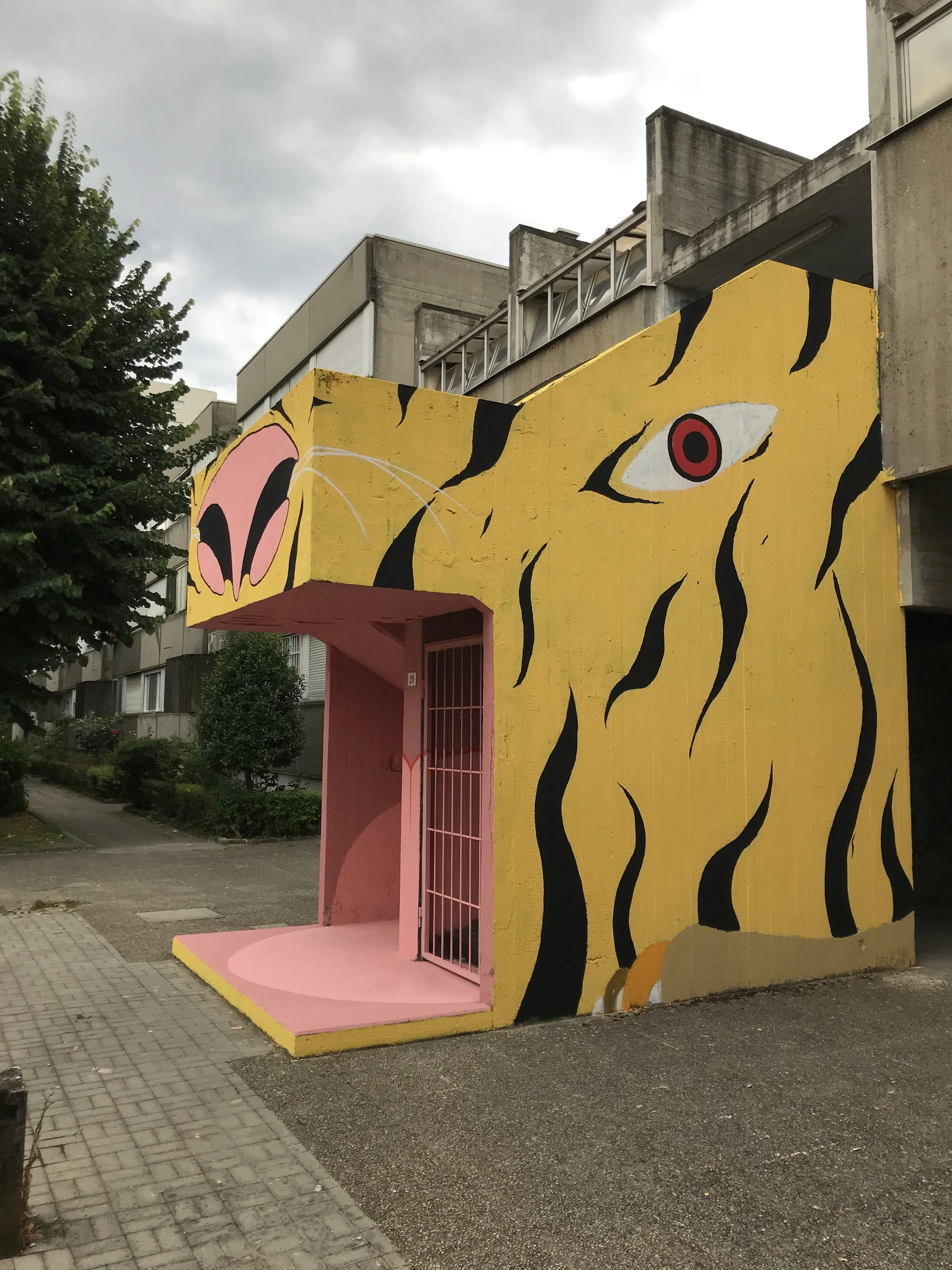 Murale colorata di un leone stilizzato su una parete gialla, con un ingresso rosa e griglia in metallo. L'edificio si trova in un'area urbana, con alberi e pavimentazione in pietra.