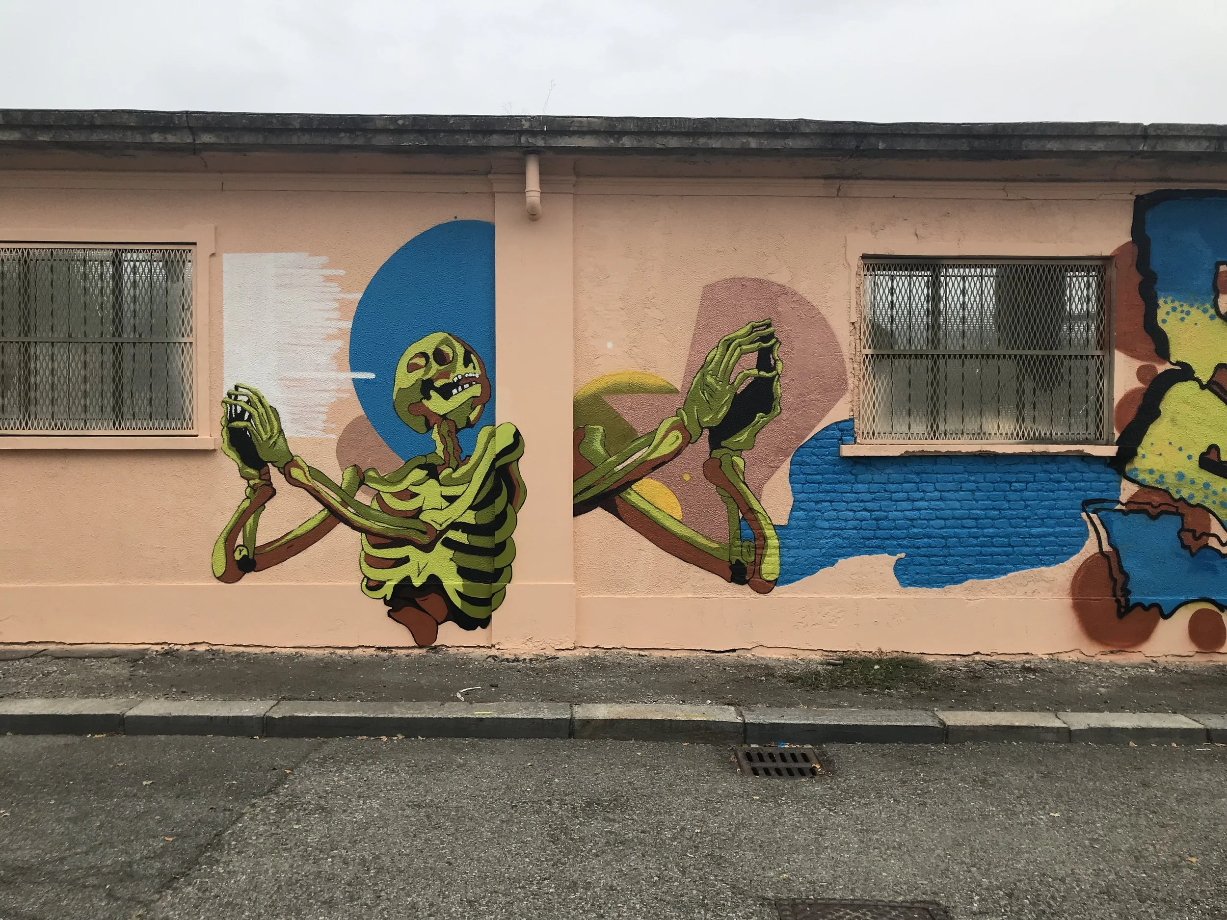 Murale colorata raffigurante uno scheletro che fa un gesto con le mani, dipinta su un muro vicino a due finestre con grate.