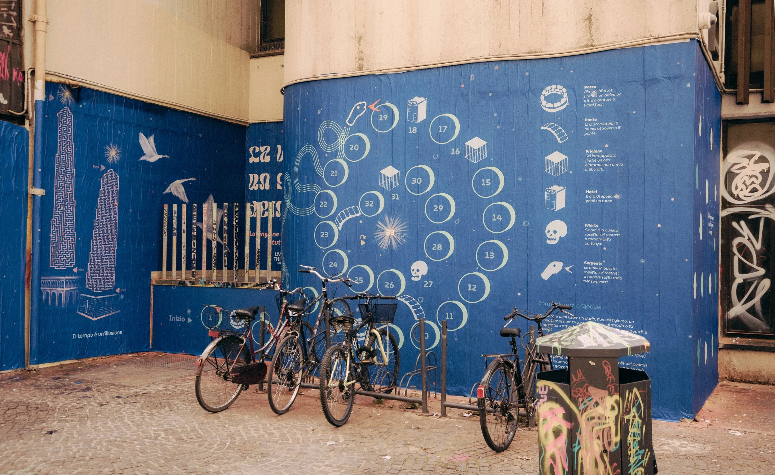 Muro blu con disegno di un gioco, che include numeri da 1 a 33, schemi e icone, con biciclette parcheggiate davanti, e graffiti su alcune pareti e un cassonetto colorato