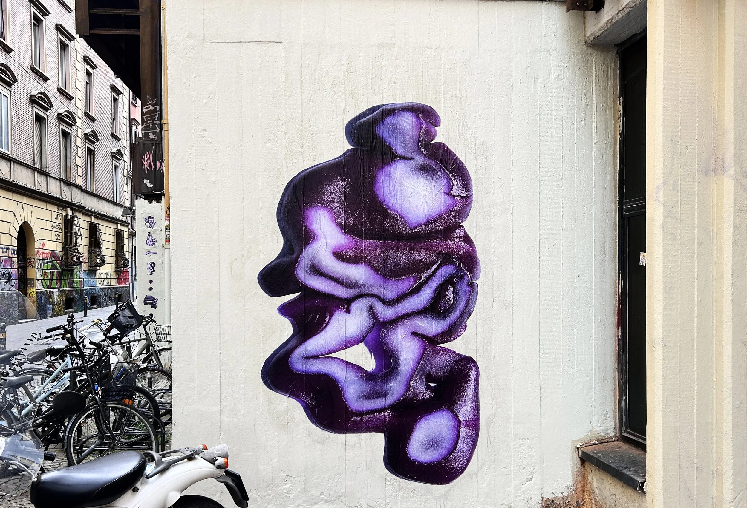Murale di graffiti che raffigura un volto umano stilizzato con tonalità di viola e bianco, posizionato su un muro bianco in un vicolo cittadino, con biciclette parcheggiate in primo piano.