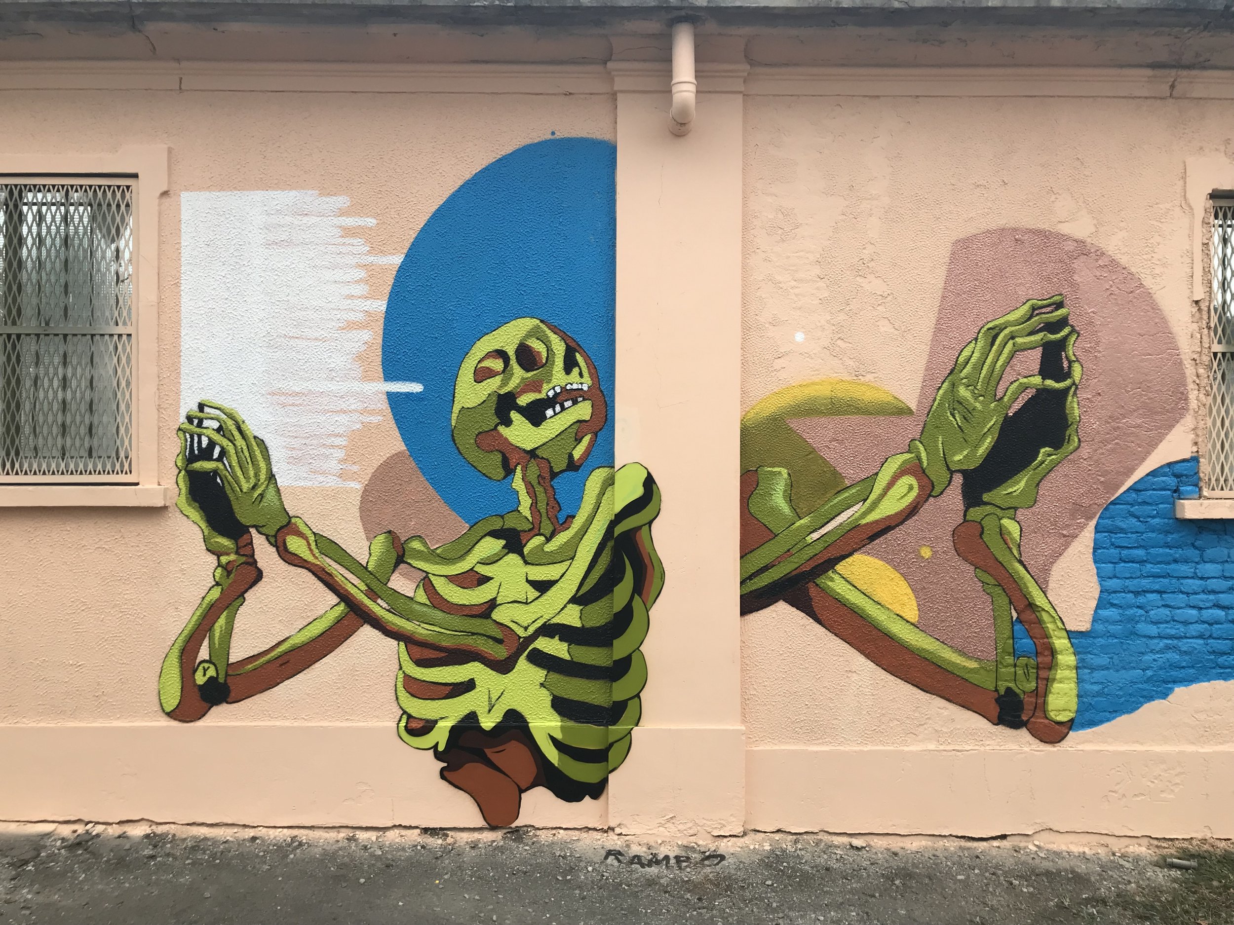 Murale colorata con disegni di un ragno verde e un volto di una creatura fantastica, con un cielo blu e elementi astratti sul muro.