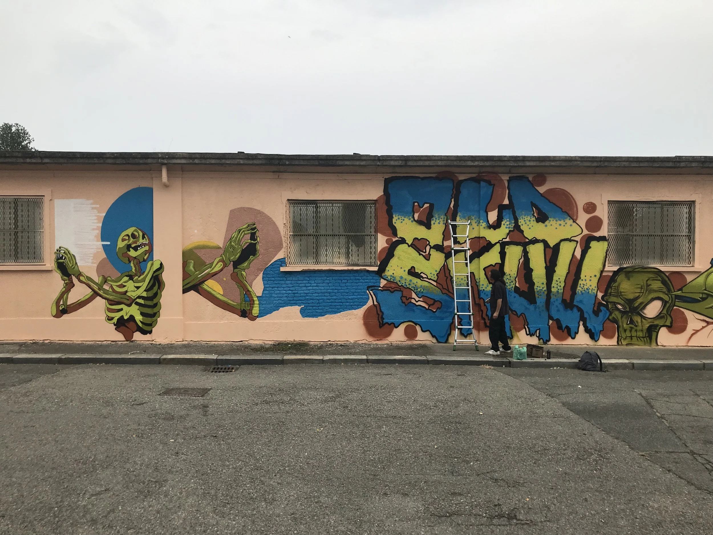 Murale colorata su un muro, raffigurante un personaggio disegnato in stile fumetto, con un volto verde e mani che prendono uno smartphone, e grandi lettere colorate.