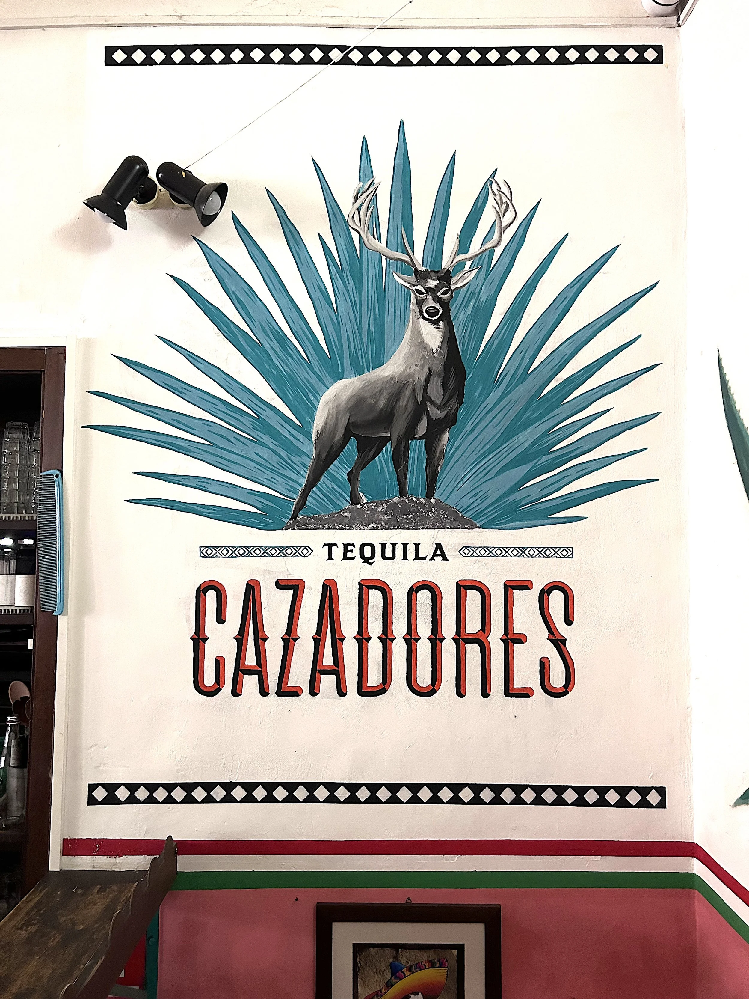 Murale colorata con un cervo a cui sono attaccate le corna, circondato da grandi foglie di colore blu. Sotto, si legge 'TEQUILA' e 'CAZADORES' in caratteri grandi e rossi, con decorazioni nere e bianche sopra e sotto.