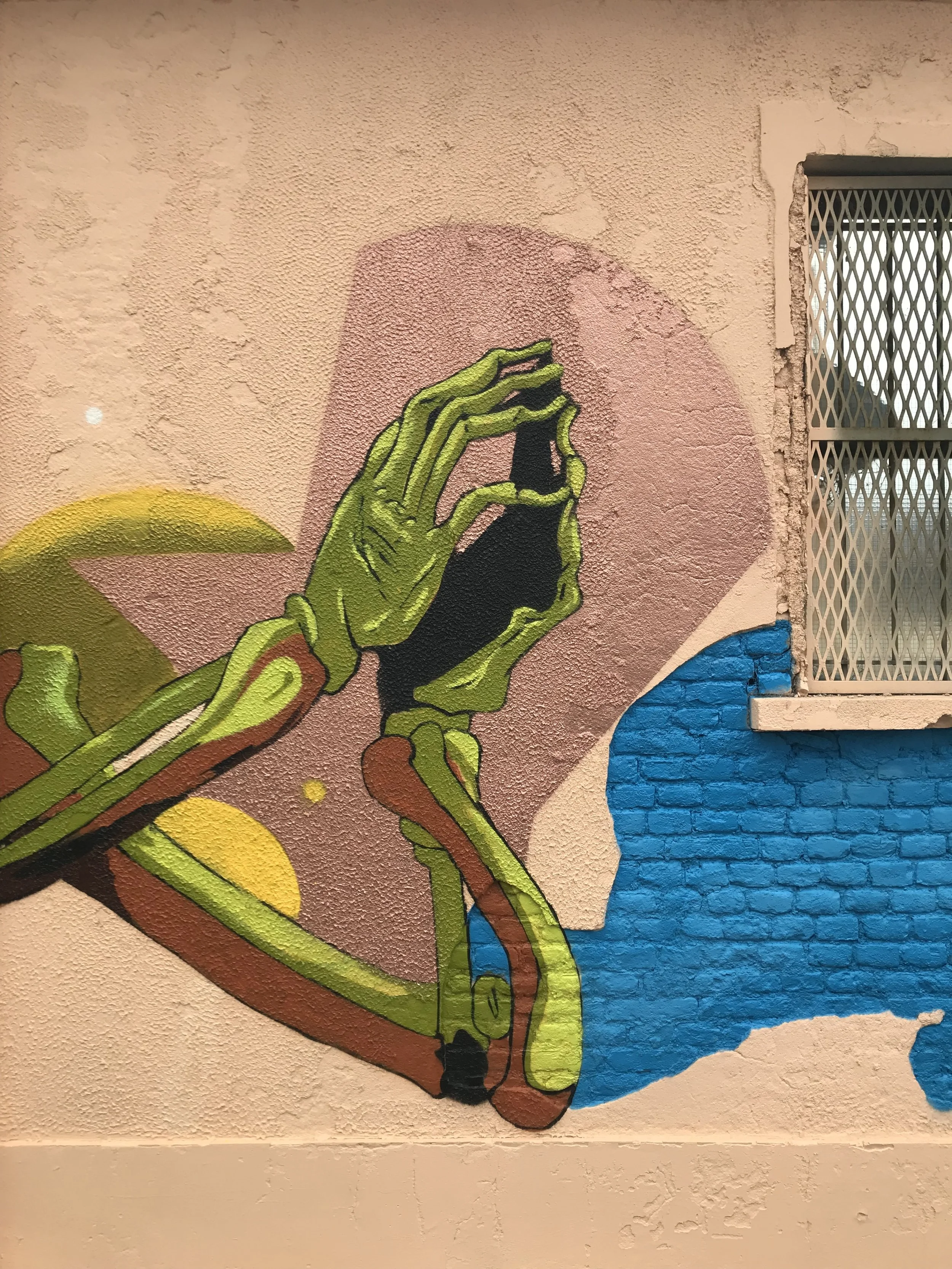 Murale street art di una mano verde che tiene uno smartphone, con sfondo colorato e dettagli di muro.