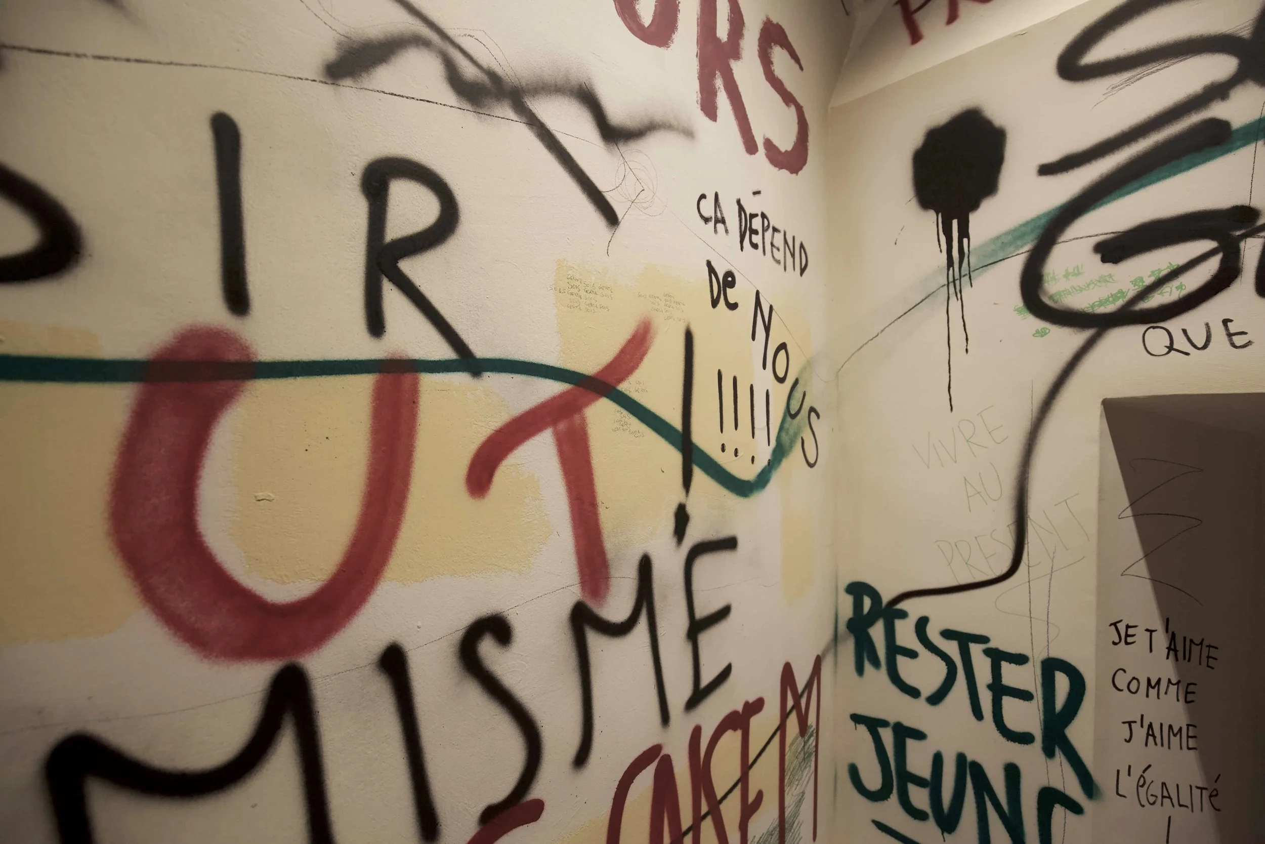 Parete coperta di graffiti e scritte colorate con vari messaggi e disegni, tra cui uno con una presenza di rosso e nero, e alcune scritte in francese.
