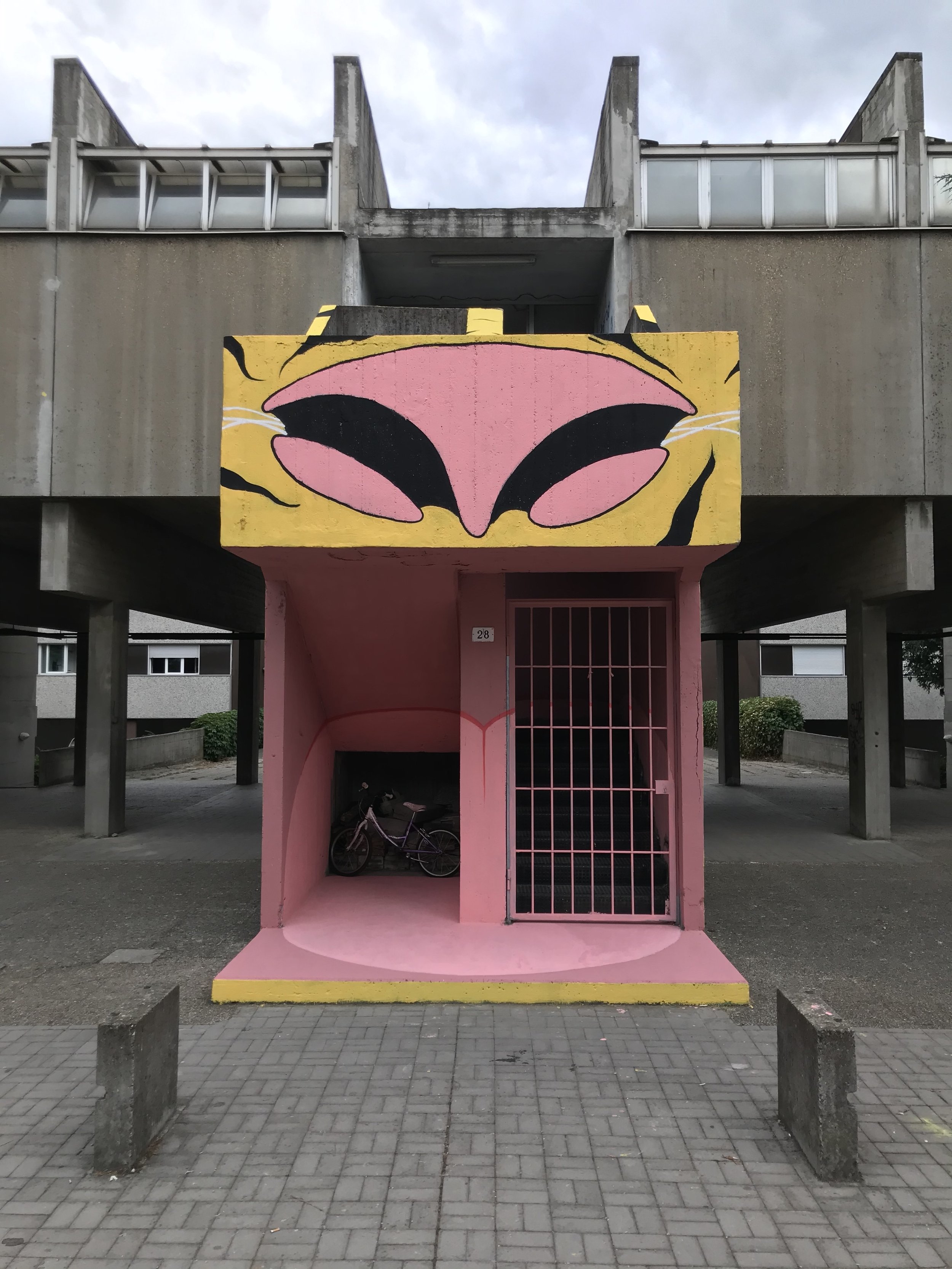Murale colorata raffigurante una bocca stilizzata con labbra rosa e lingua nera su una struttura rosa con dettagli gialli.