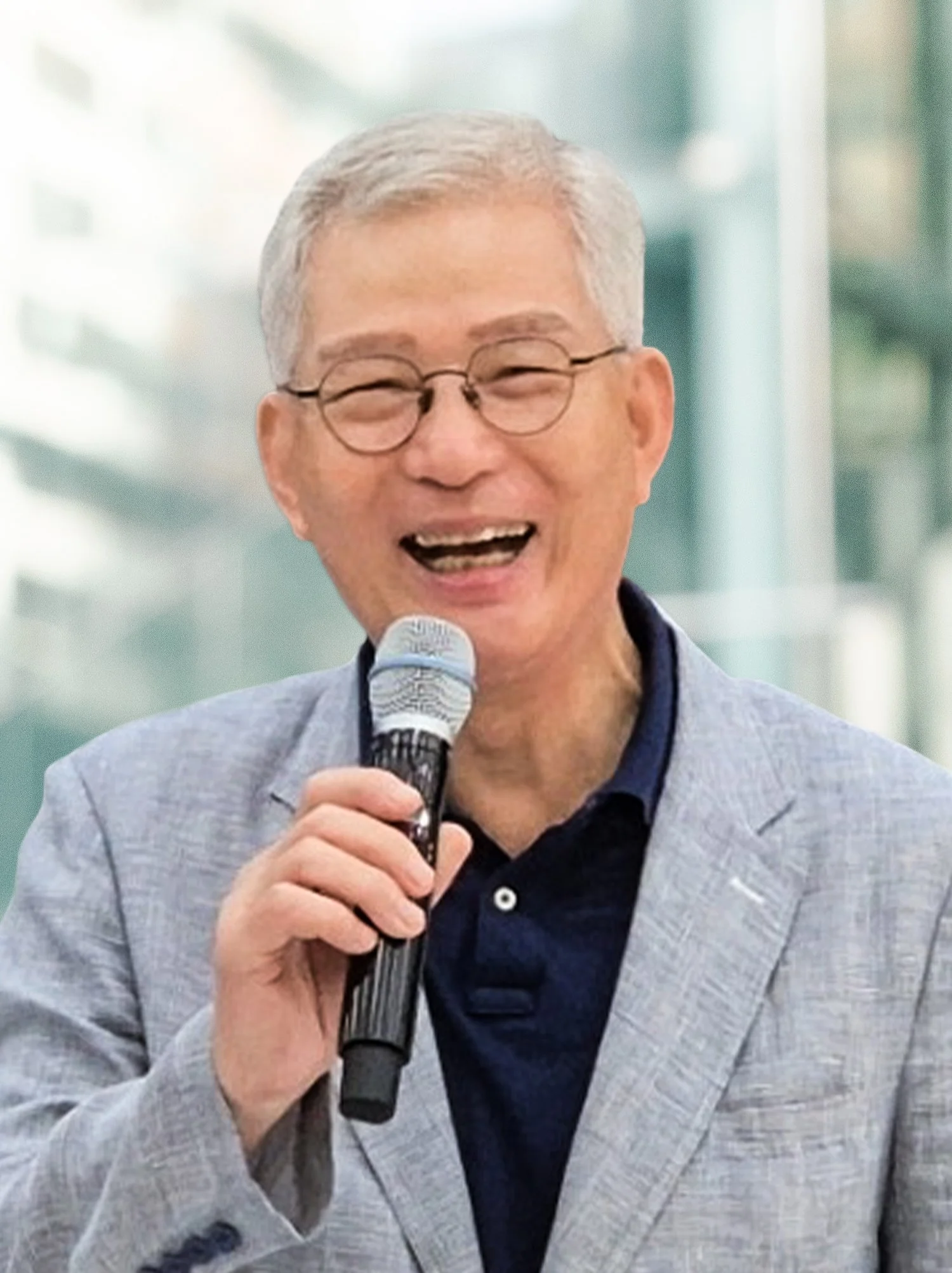 A smiling elderly man with glasses and gray hair, holding a microphone and speaking at an event, dressed in a light gray blazer and a dark shirt, with a blurred modern office building in the background.