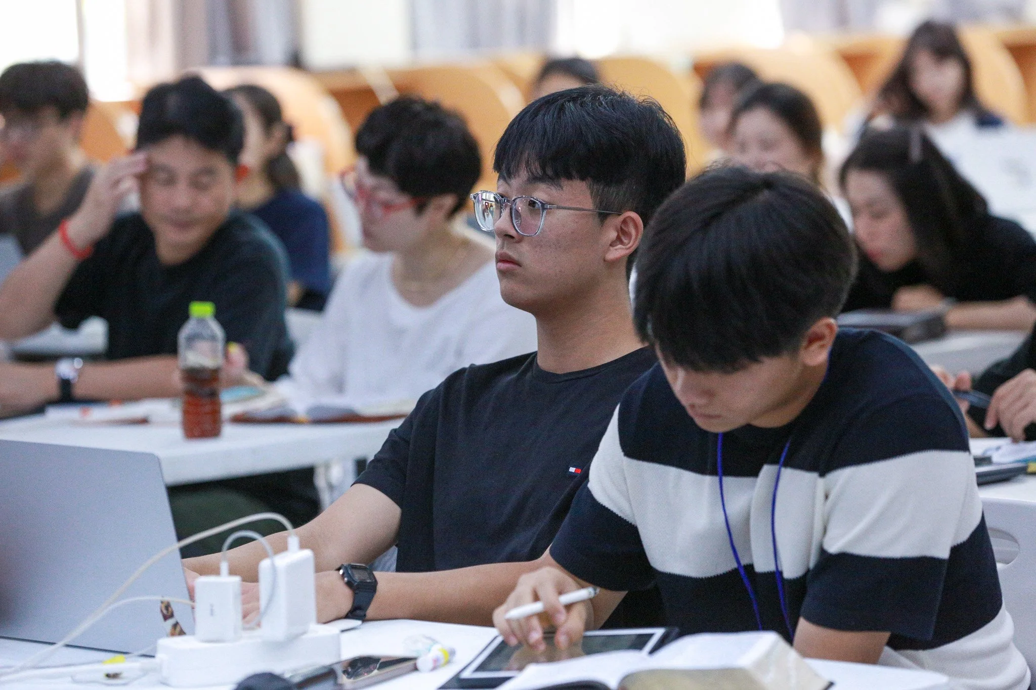 Students attending a lecture in a classroom, taking notes and working on laptops.