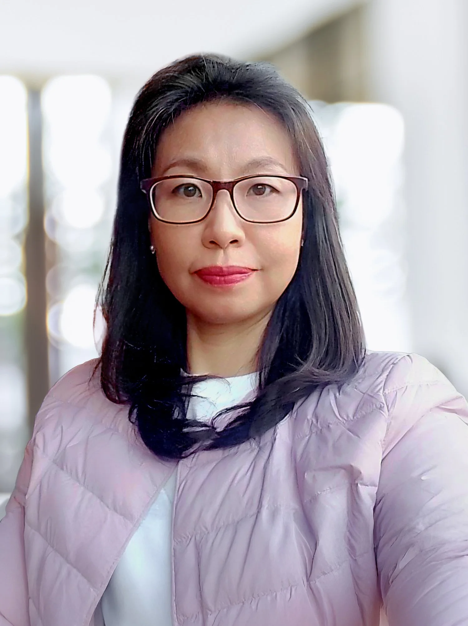 A woman with long black hair, glasses, and wearing a light pink quilted jacket, taking a selfie in a bright, blurred indoor setting.