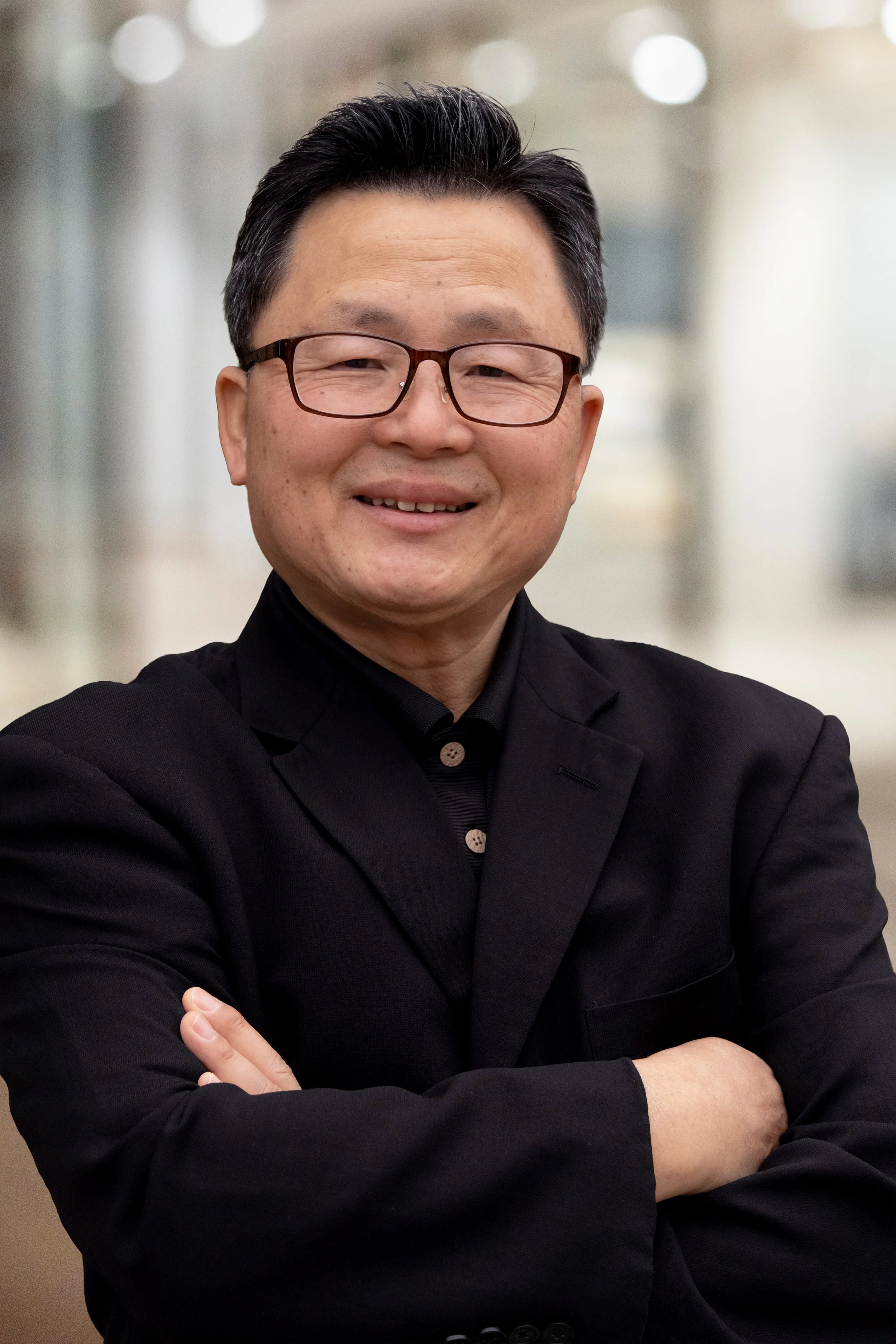 A man smiling with glasses, wearing a black blazer and shirt, crossing his arms in an indoor setting with blurred background.