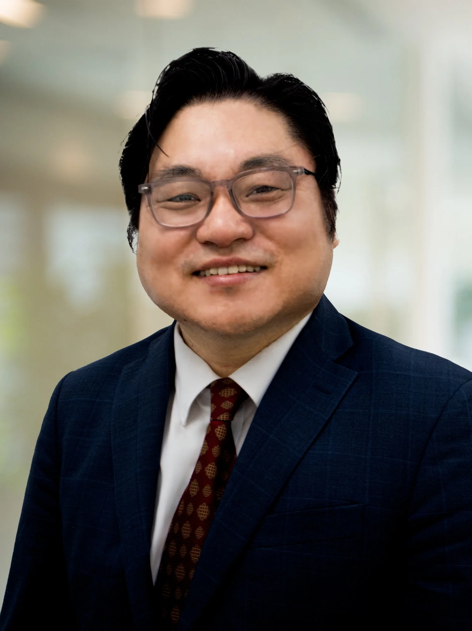 A man wearing glasses, a navy blue suit, white shirt, and patterned brown tie, smiling with a blurred indoor background.