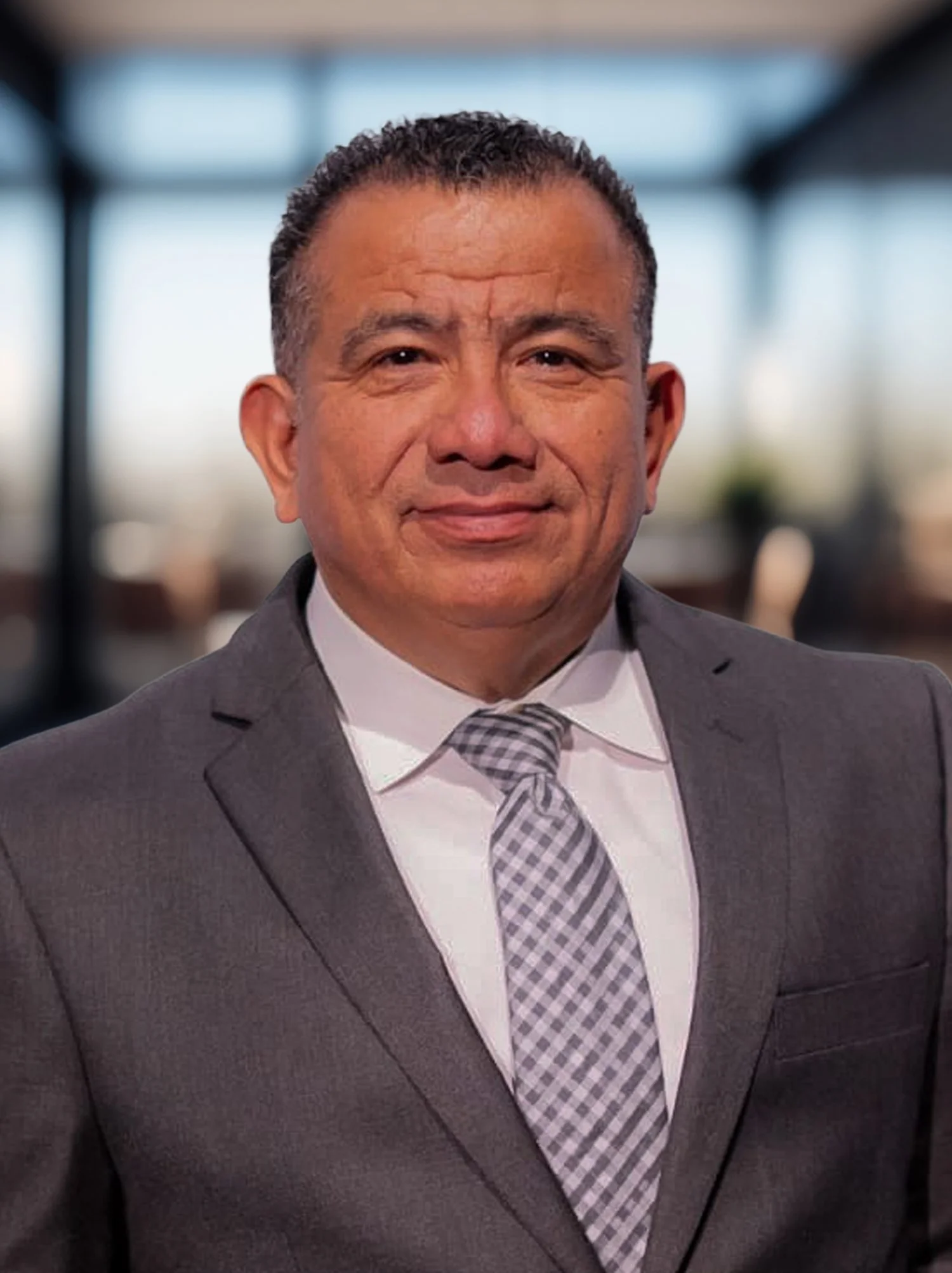 A middle-aged man in a gray suit, white shirt, and checkered tie, smiling in an indoor setting with blurred background.