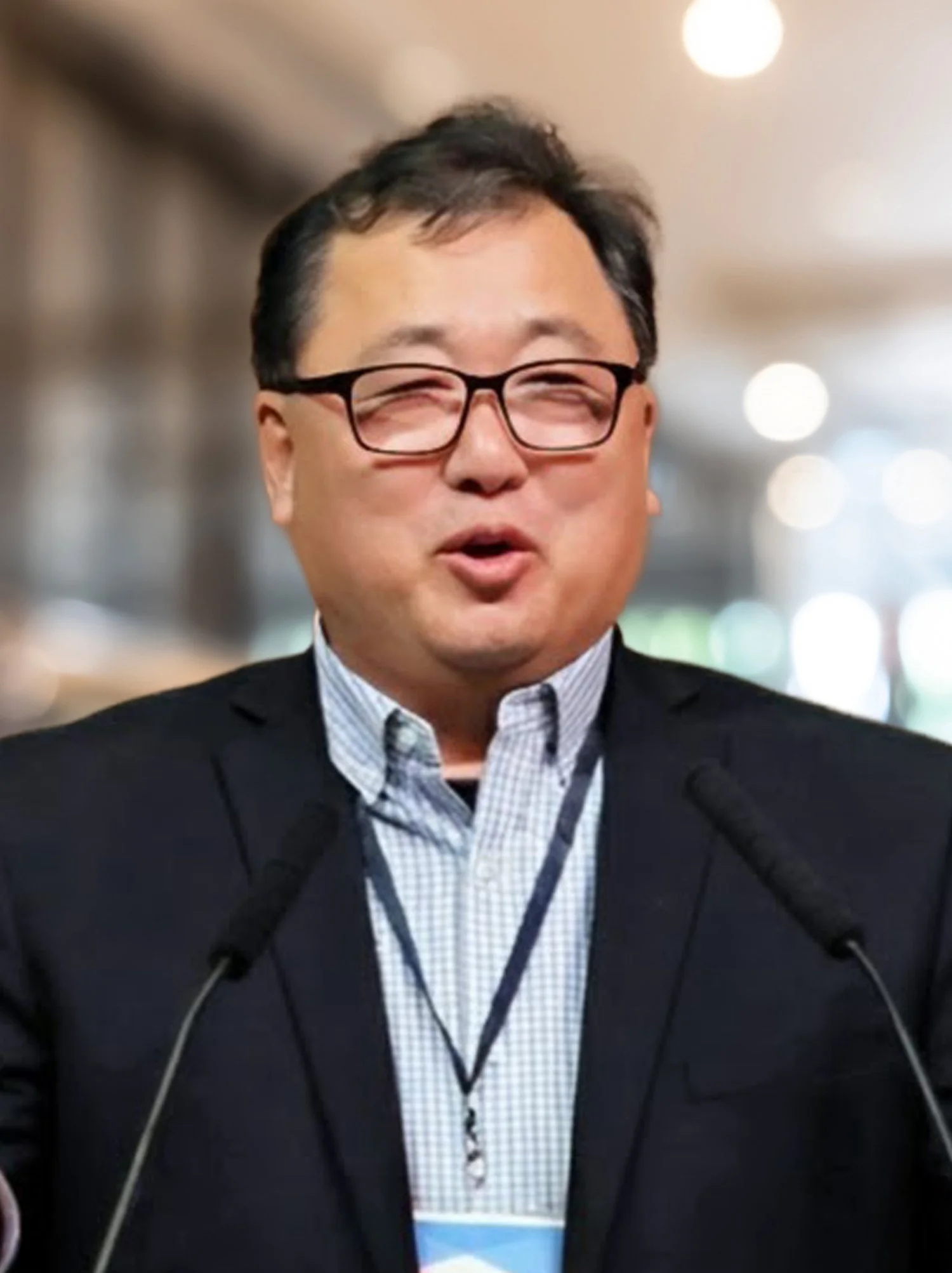 A man with glasses speaking at a podium with microphones in a professional setting.