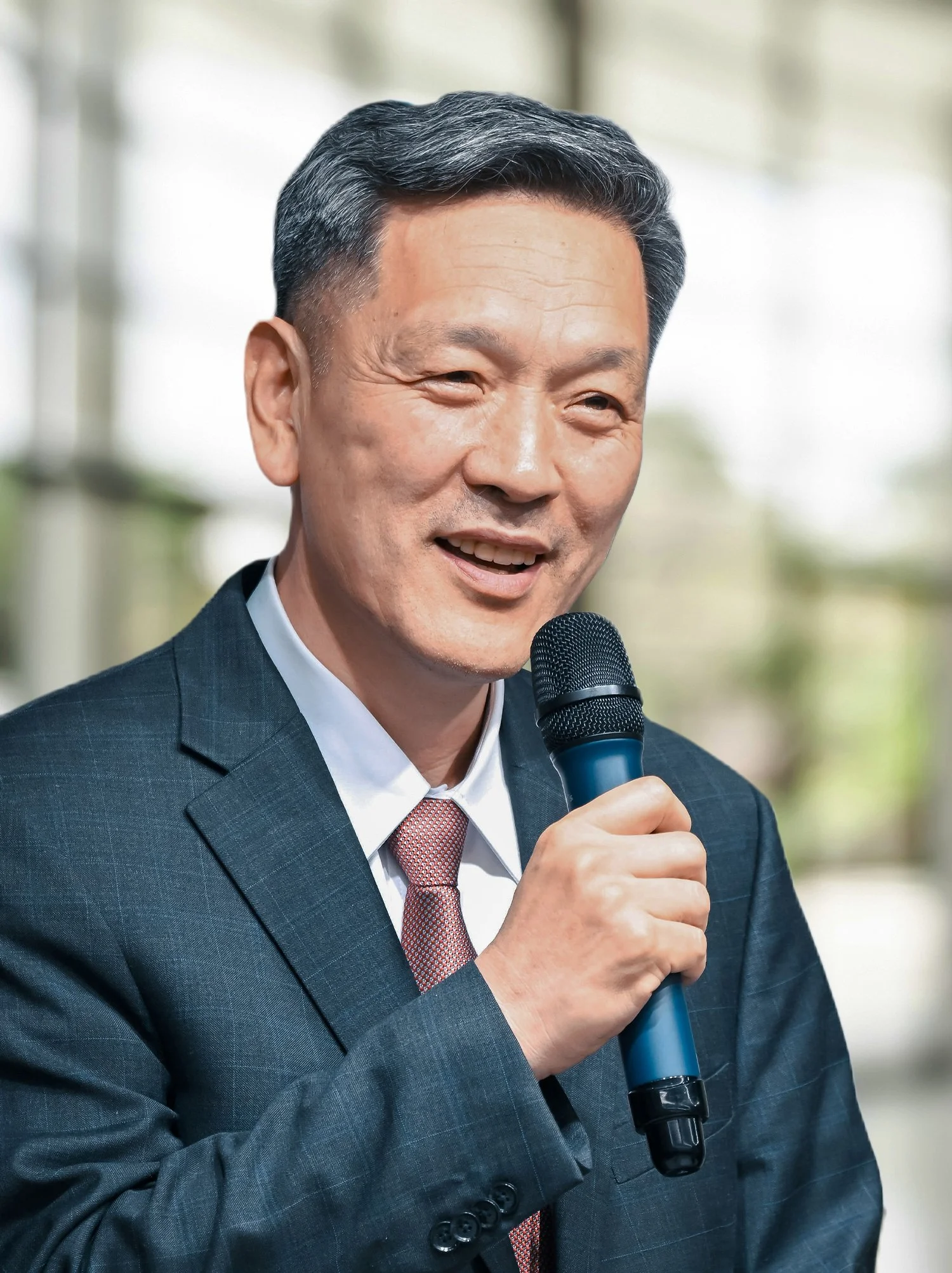 A middle-aged Asian man in a business suit holding a microphone and smiling during a speech or presentation.