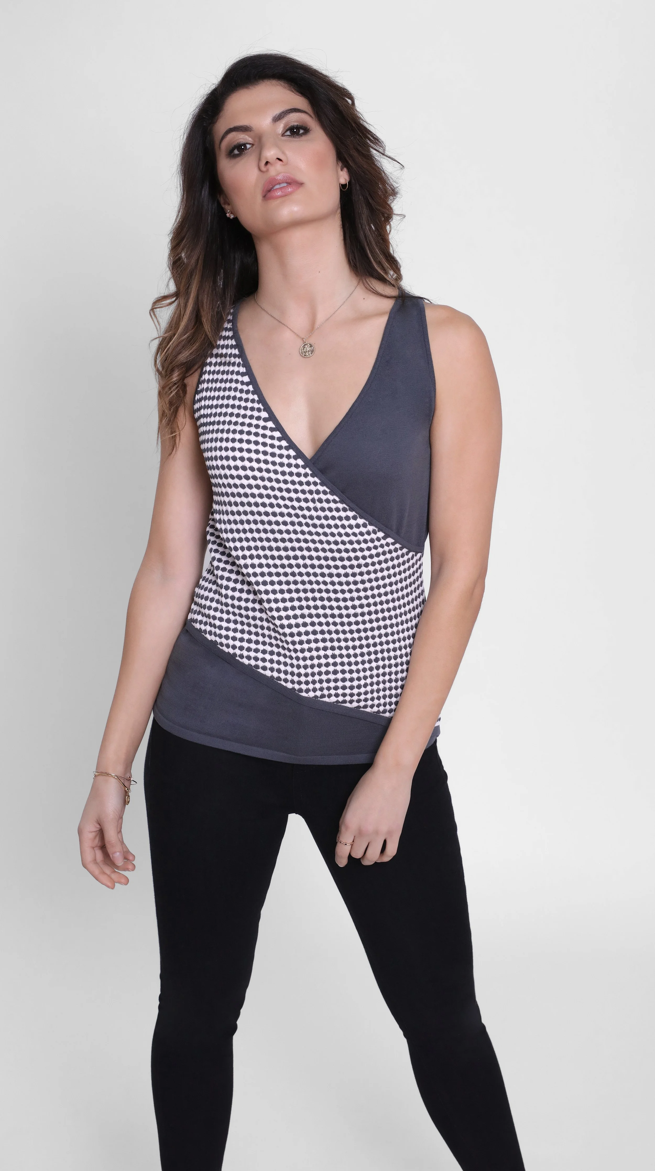 Woman with wavy brown hair wearing a sleeveless top with a black and white mesh pattern and black pants, standing against a plain white background.