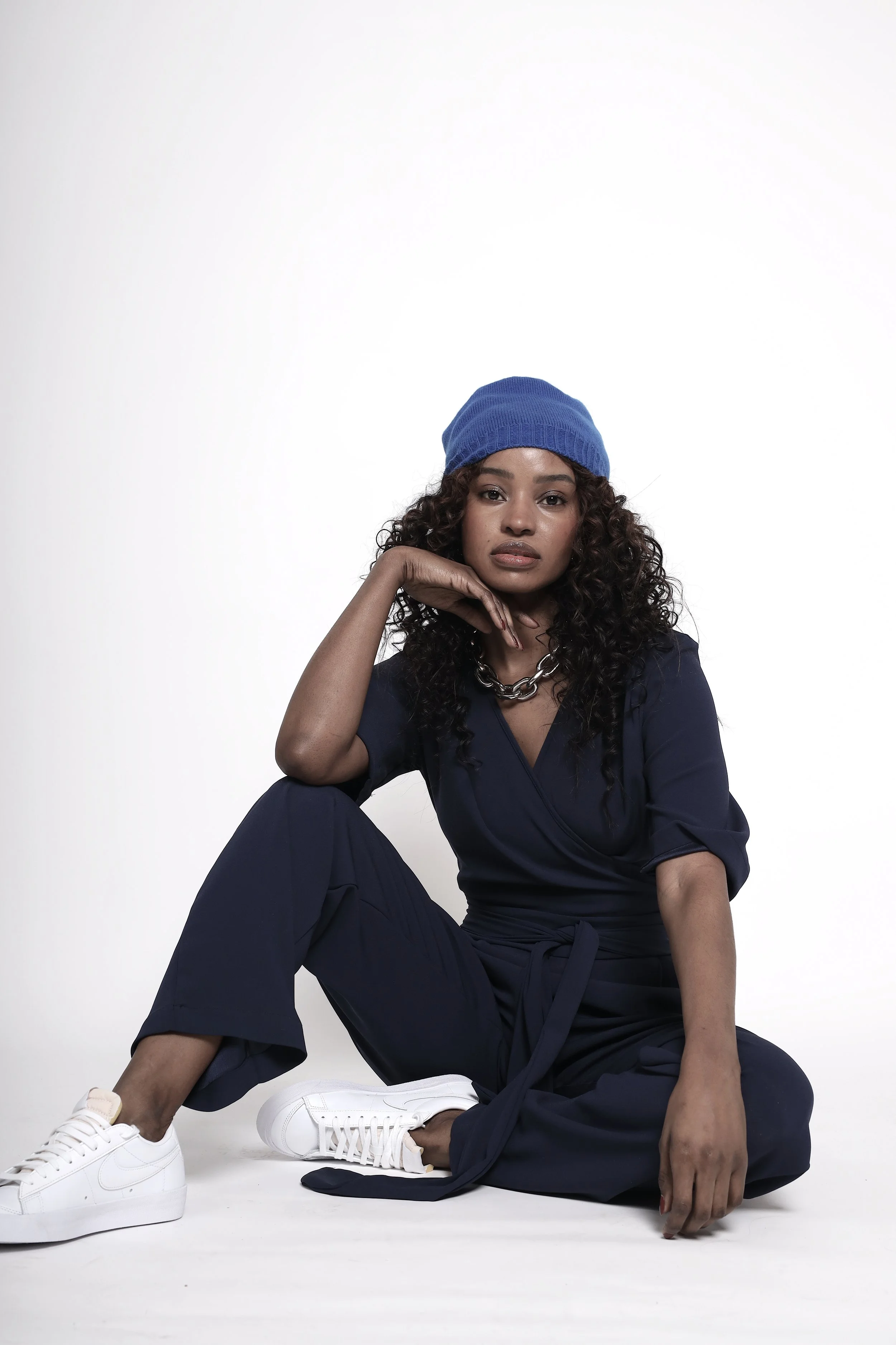 A woman with curly hair wearing a blue beanie, navy jumpsuit, and white sneakers, sitting on the floor against a plain white background.
