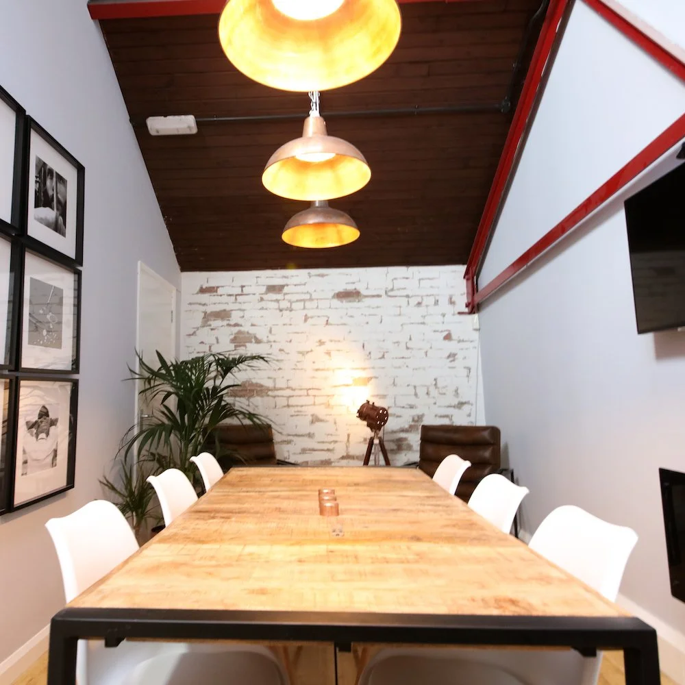 A modern conference room with a wooden table, white and black chairs, a white brick wall, and hanging pendant lights. There is a large plant and a vintage camera on a tripod.