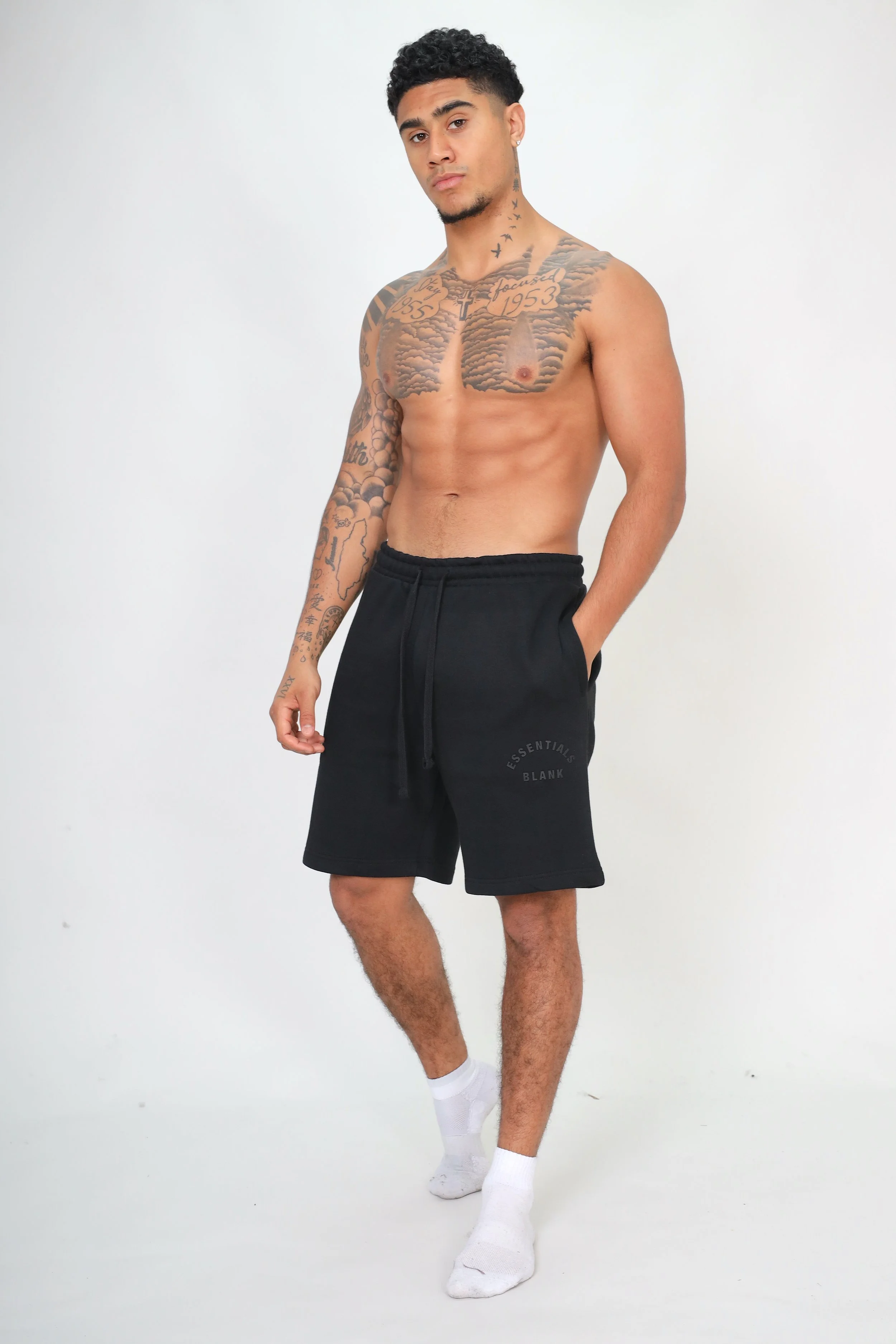 A young man with tattoos, wearing black shorts and white socks, standing against a plain white background.