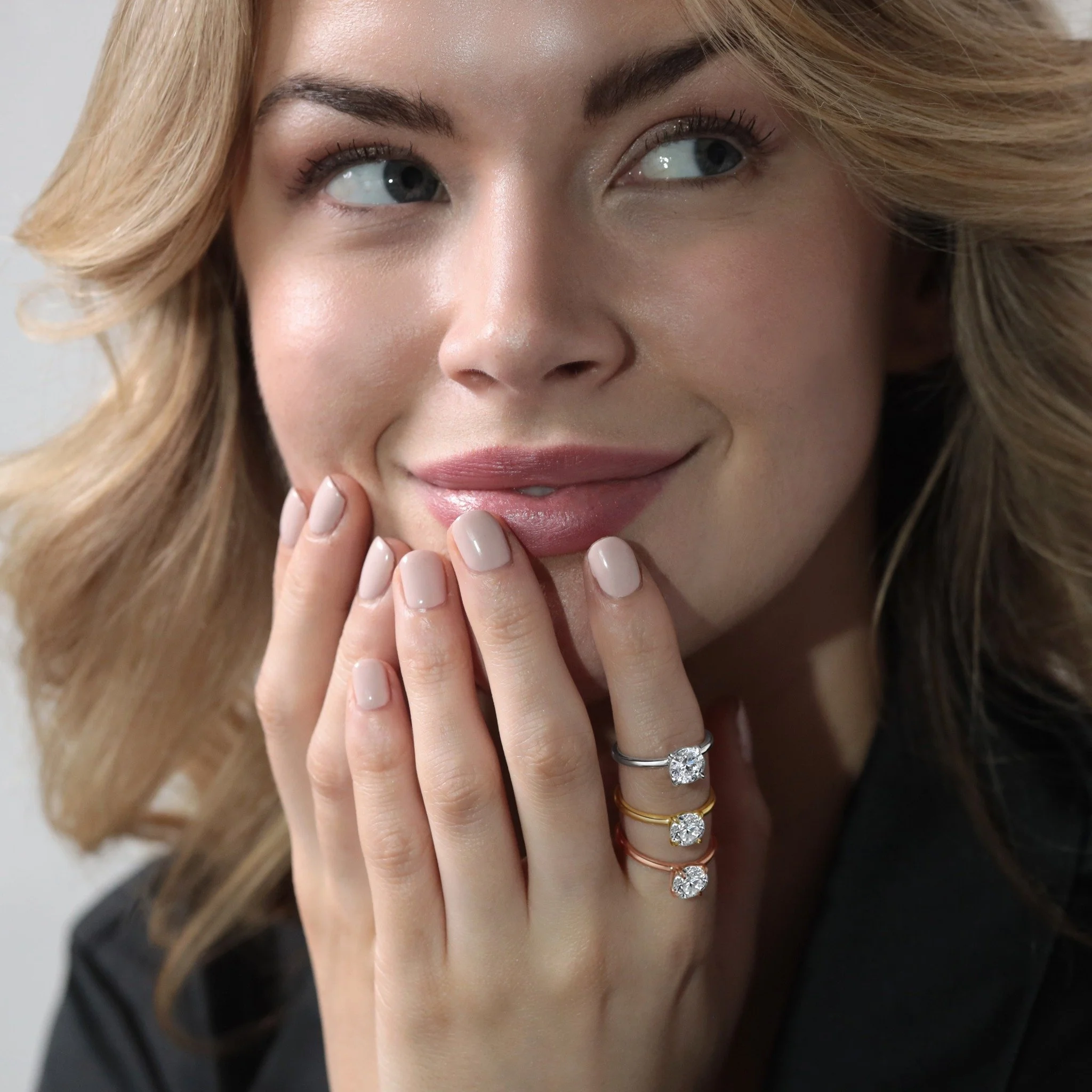 A woman with blonde hair and blue eyes gently touching her face with her hand, wearing multiple rings with diamonds and a nude lip color.