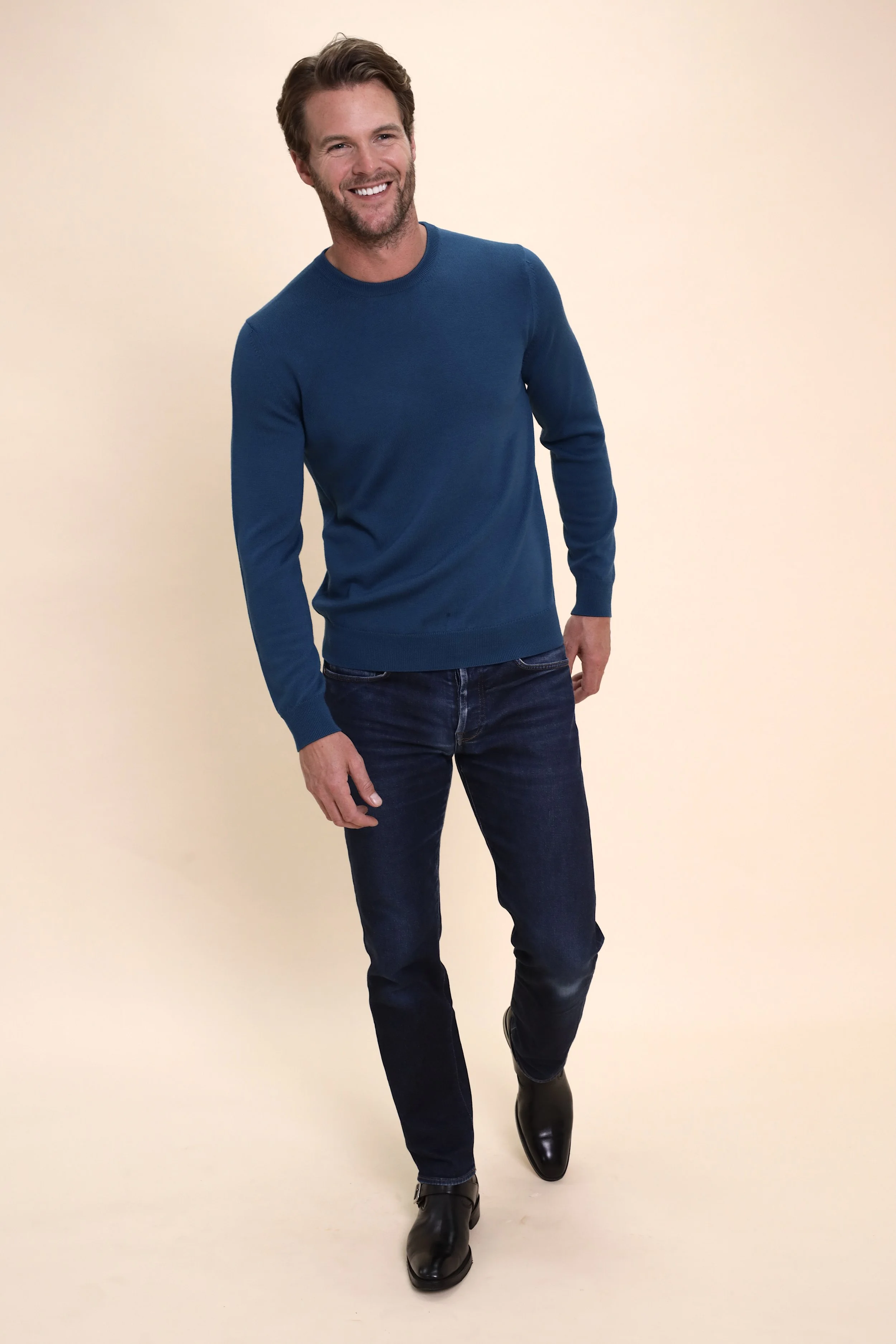 Smiling man in a blue sweater, dark jeans, and black boots standing against a beige background.