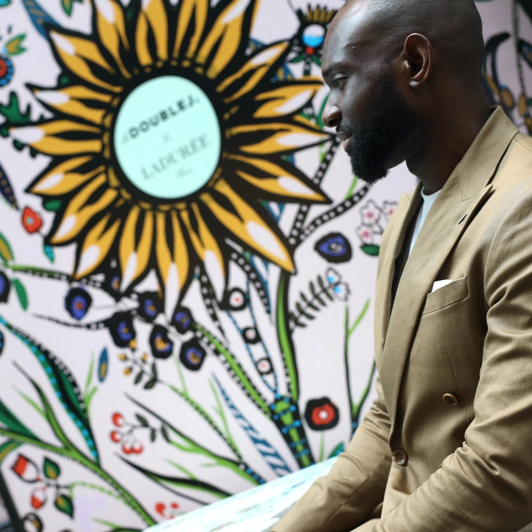 A man dressed in a beige blazer with a white pocket square, sitting and looking thoughtfully downward. In the background, there is a colorful, artistic mural featuring a large sunflower with the words 'Double x Laughter' inside the flower's center, s