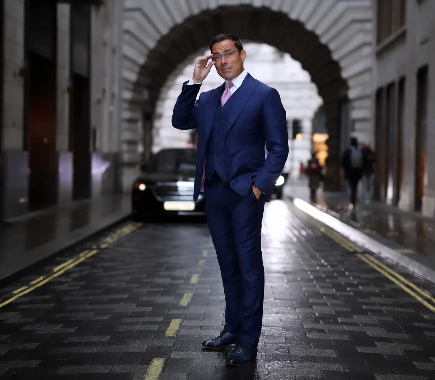 A well-dressed man in a blue suit, purple tie, and glasses adjusts his glasses while standing on a wet city street with an archway in the background and a black luxury car behind him.