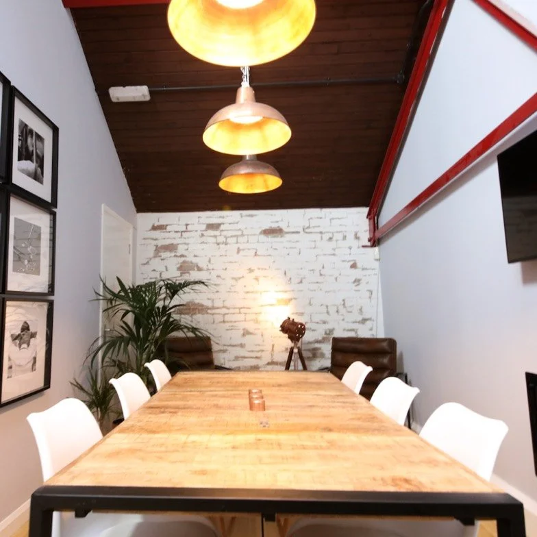 A modern conference room with a long wooden table, white chairs, a white brick accent wall, framed black-and-white photos, a potted plant, and overhead pendant lights.