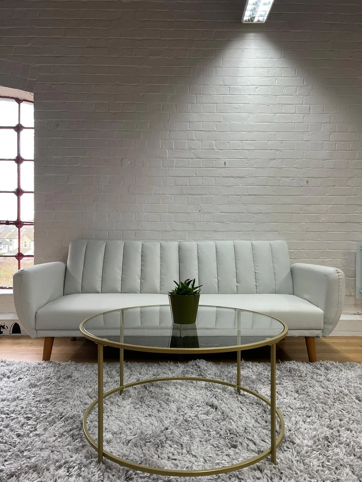Modern living room with a white leather sofa, a gold and glass coffee table with a small green potted plant, a white fluffy rug, a brick wall, and a window with red grid design.