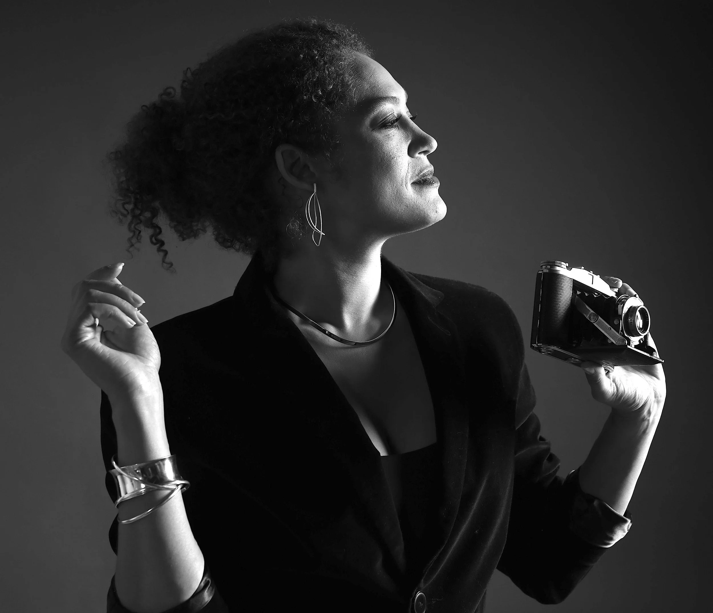 Black and white portrait of a woman with curly hair holding a vintage camera, wearing earrings, a necklace, and a bracelet, with a confident expression.