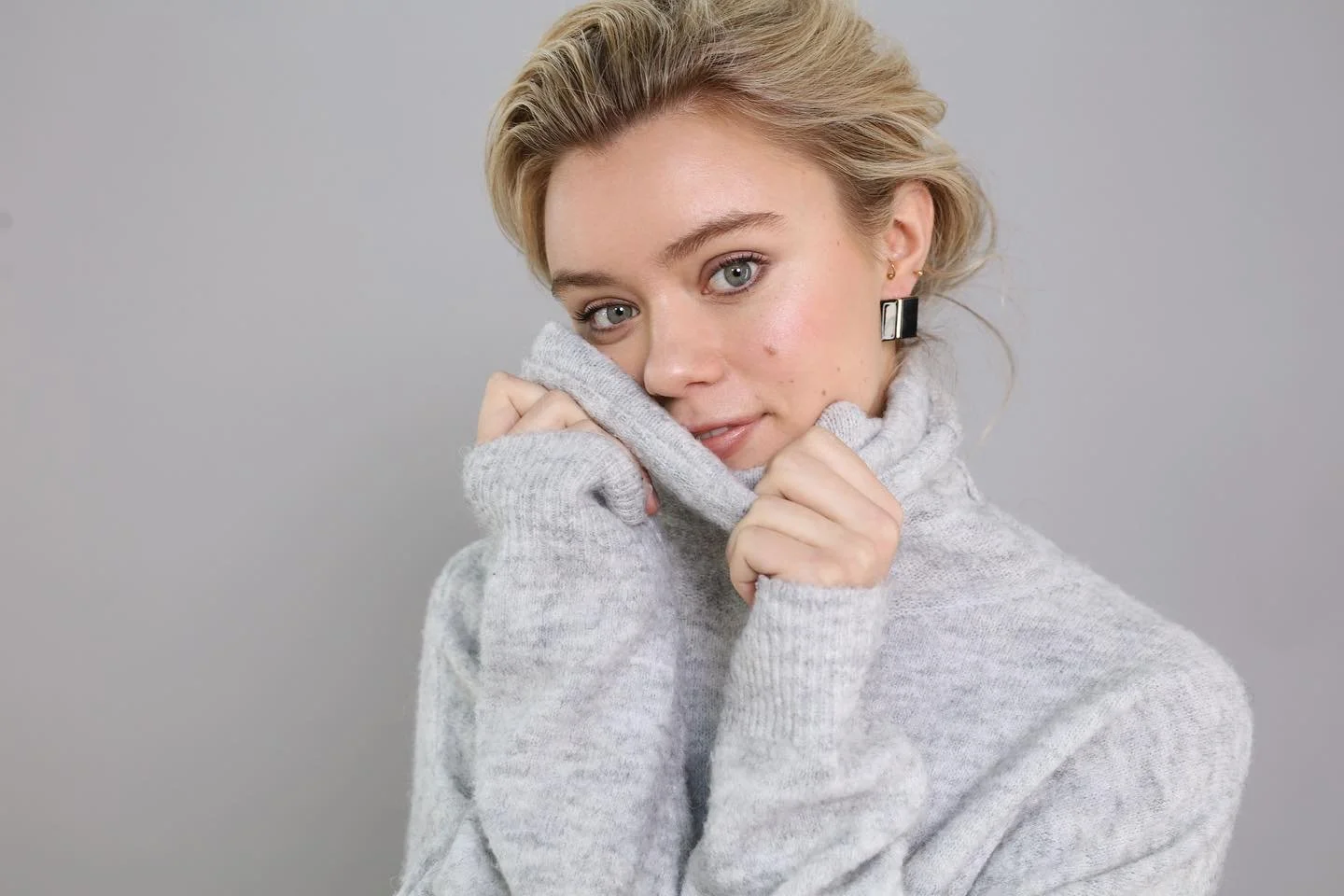 A woman with blonde hair, wearing a light grey sweater and black earrings, looks directly at the camera, holding the high collar of her sweater up to her face against a plain grey background.