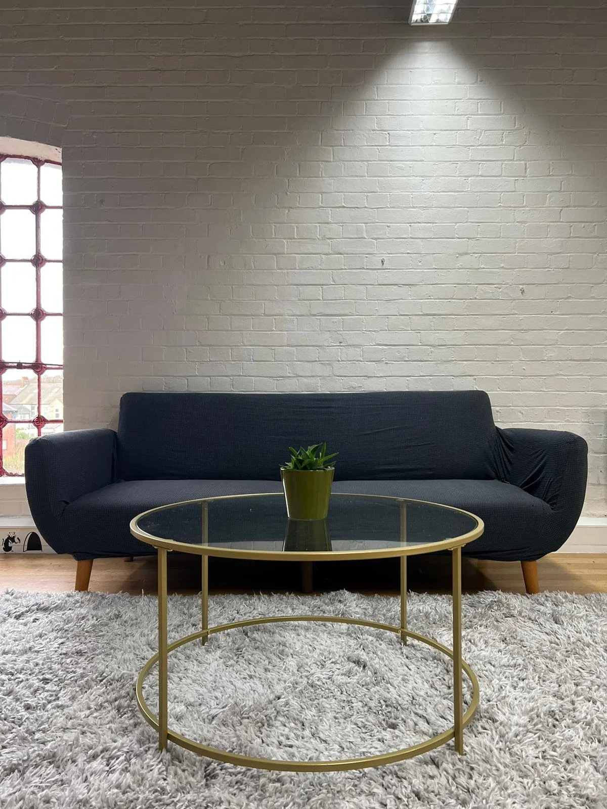 Living room with a dark blue sofa, a glass-top coffee table with a gold frame, a green potted plant on the table, a white brick wall, a window with a grid pattern, and a fluffy white rug.