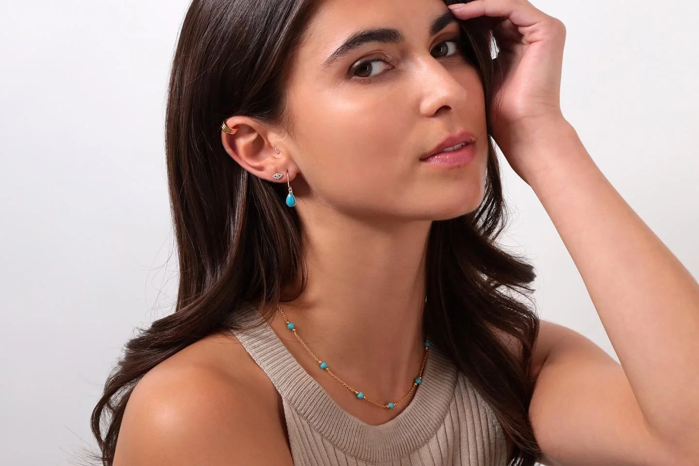 A woman with brown hair and light skin wearing turquoise jewelry, including earrings, a necklace, and a top with a beige strap, posed with her hand near her head against a plain white background.