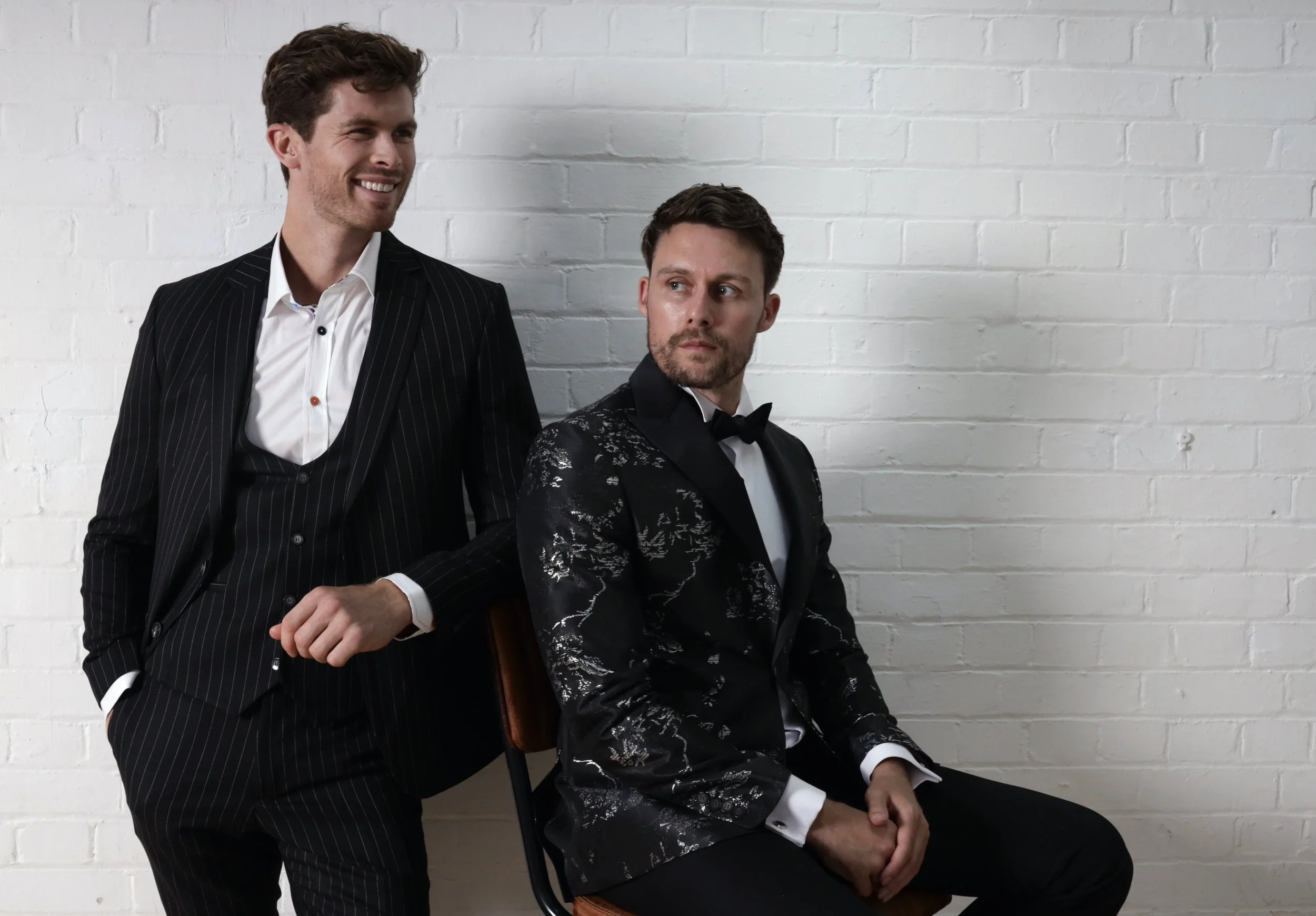Two men in tuxedos, one sitting and one standing, against a white brick wall. The man standing is smiling, while the seated man looks to the side.