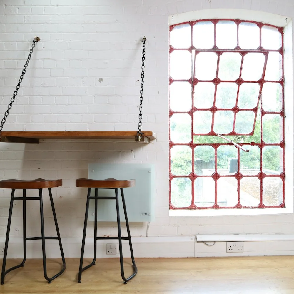 Interior view of a room with a white brick wall, a large window with red metal framing, a floating wooden shelf suspended by chains, two wooden bar stools with black metal legs, and electrical outlets on the wall.
