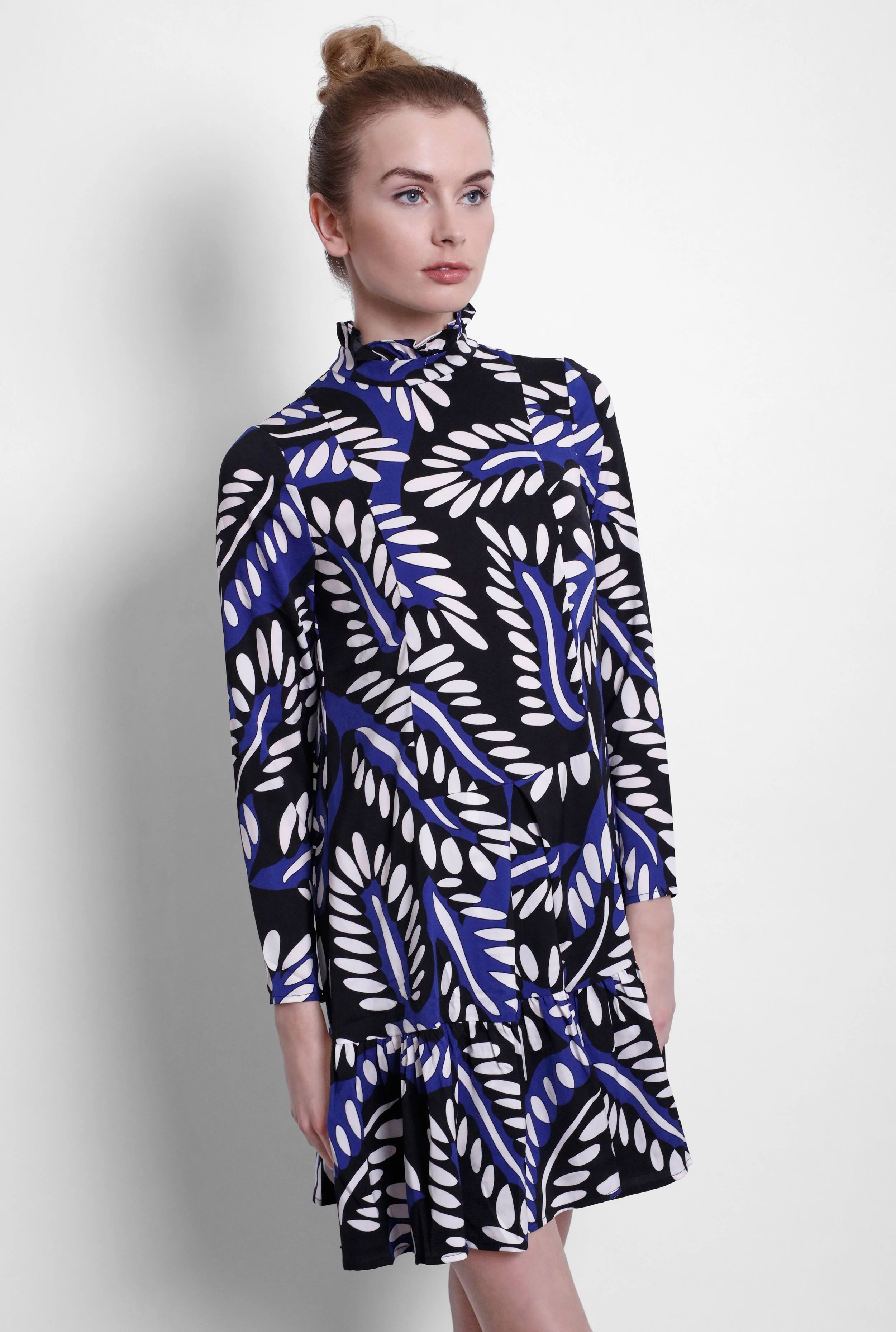 A woman with fair skin and red hair tied in a bun, wearing a black, blue, and white patterned dress with a ruffled collar, standing against a plain white background.