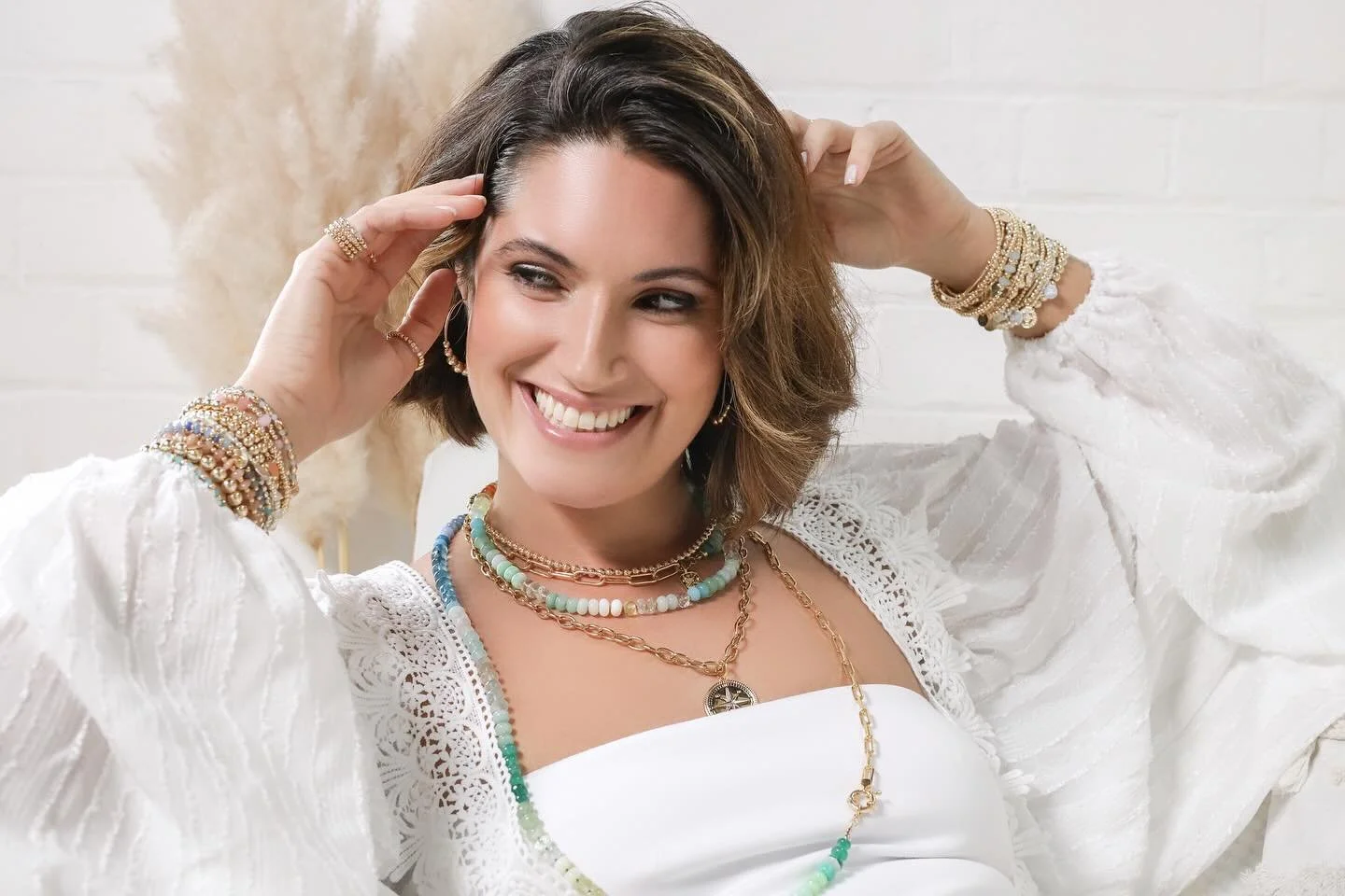 A woman with brown hair smiling, wearing multiple colorful jewelry pieces including necklaces, earrings, and bracelets, dressed in a white, textured top with lace details, sitting against a white brick wall.