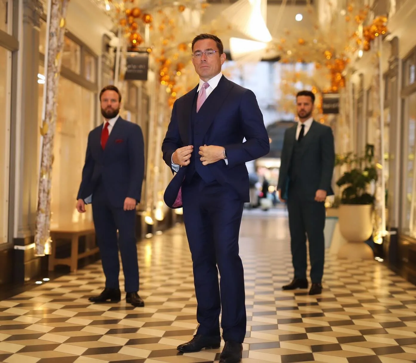 Three men in suits standing in an elegant, decorated corridor with patterned floor tiles and holiday ornaments hanging from the ceiling.