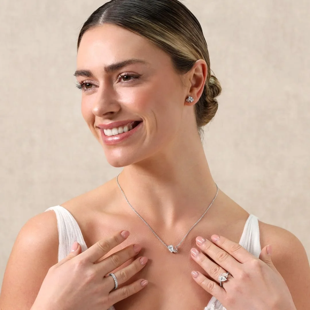 A woman with a bun hairstyle wearing diamond earrings, a diamond necklace, and a matching ring, smiling and touching her necklace.