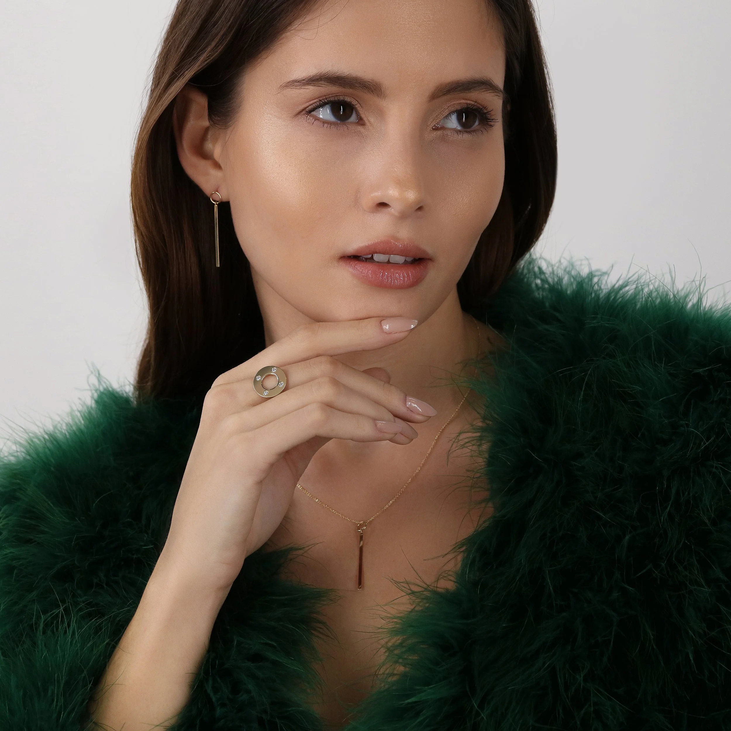A woman with long brown hair wearing gold jewelry, including a ring, earrings, and a necklace, dressed in a green fluffy coat, looking to the side with her hand near her chin.
