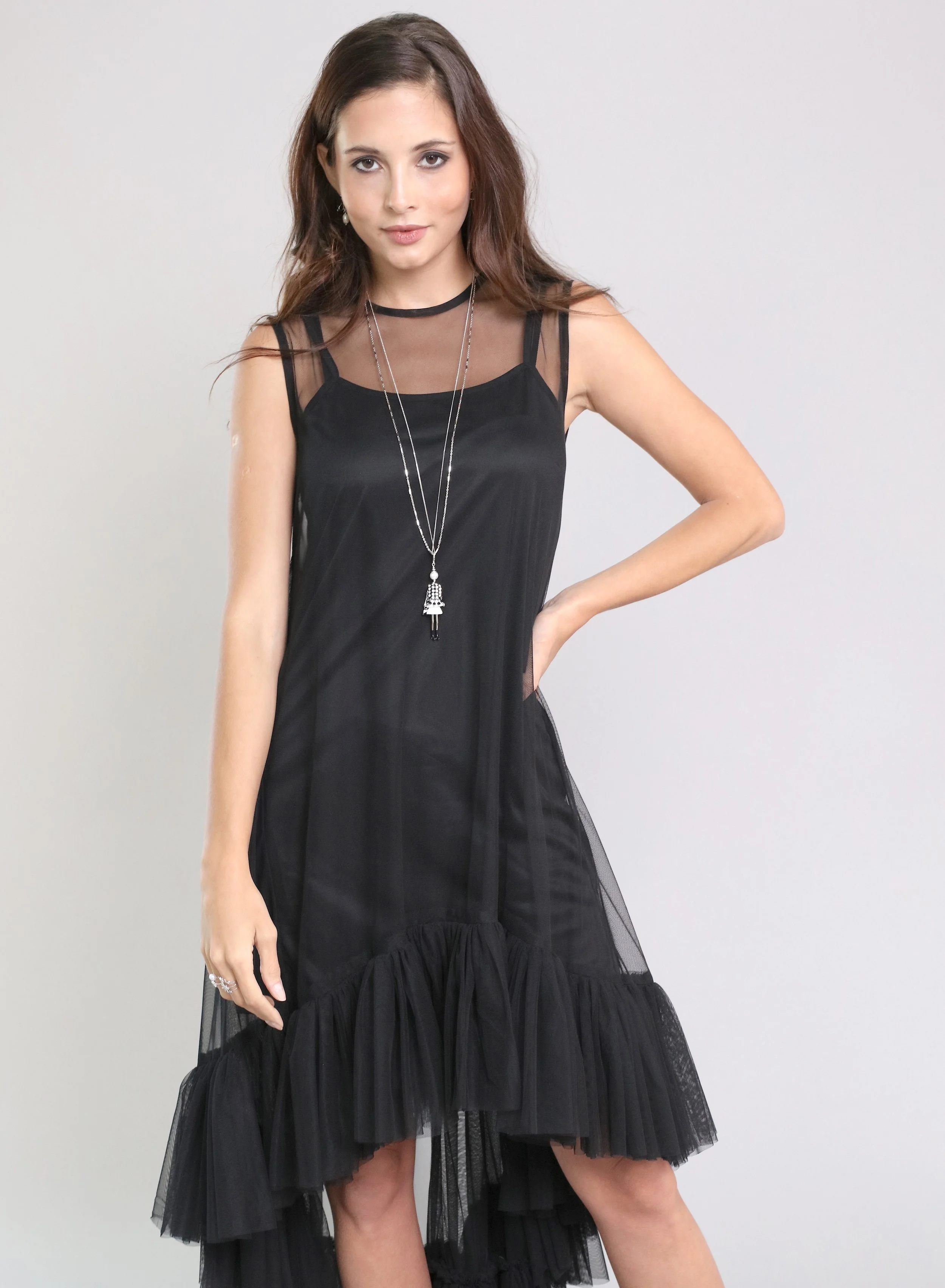 A young woman with long brown hair wearing a black sleeveless dress with sheer details and ruffled bottom, accessorized with layered silver necklaces and earrings, posing with her left hand on her hip against a plain background.