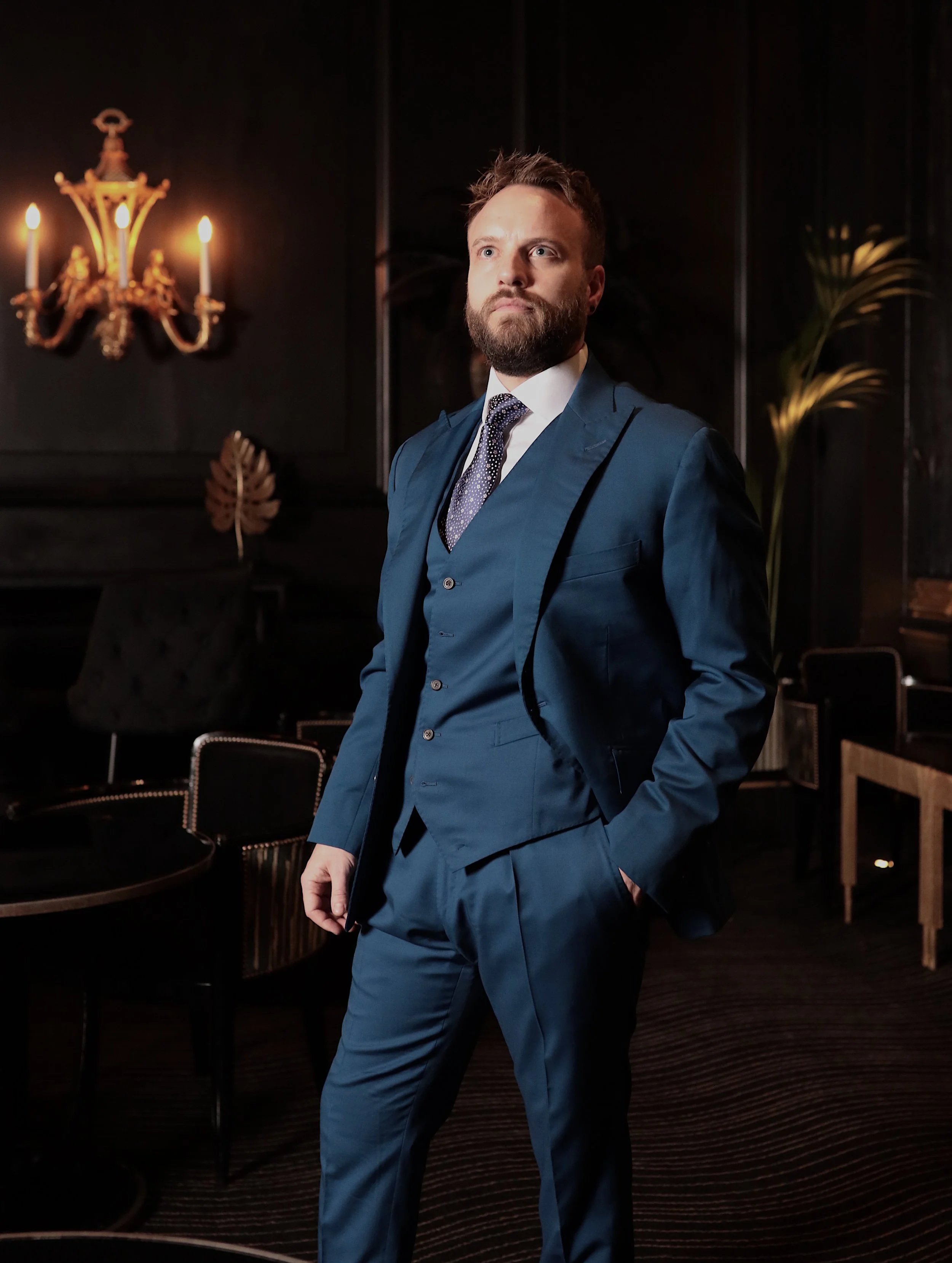 A man in a blue suit standing in a dimly lit room with dark walls and a chandelier.