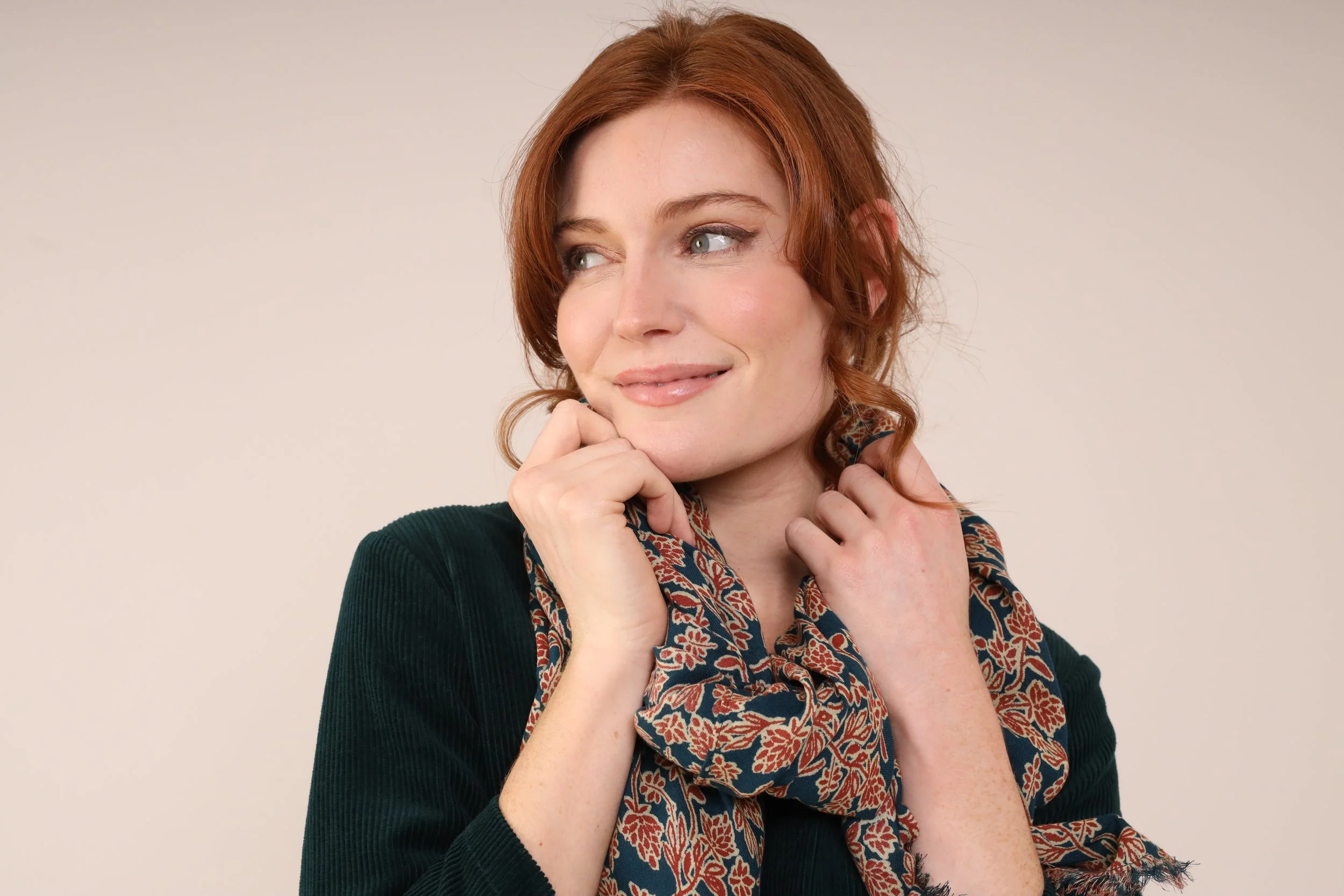 A woman with red hair smiling gently, gently touching her chin with one hand, wearing a dark top and a patterned scarf.