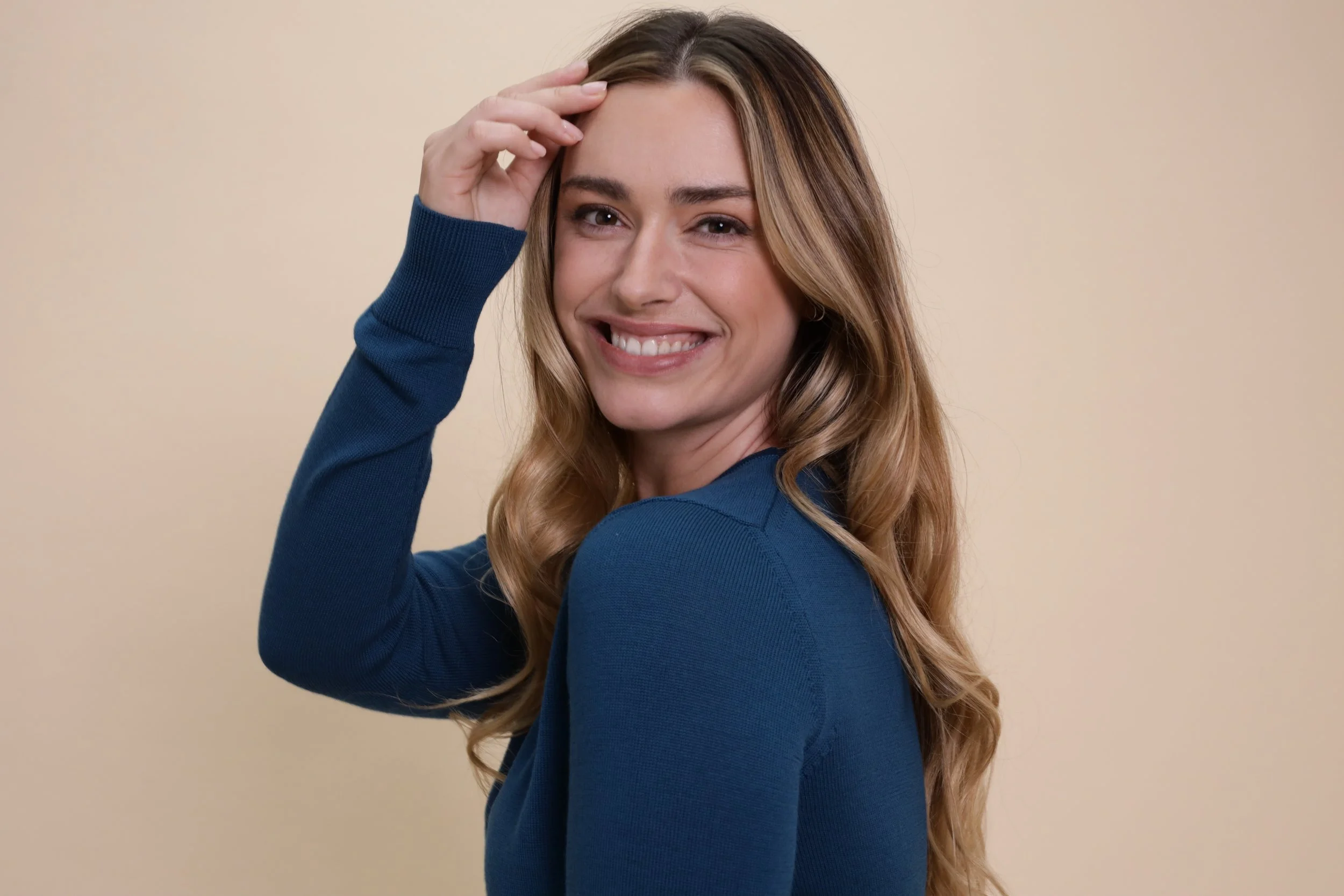 A woman with long wavy blonde hair smiling and touching her forehead, wearing a blue long-sleeved top, against a plain beige background.