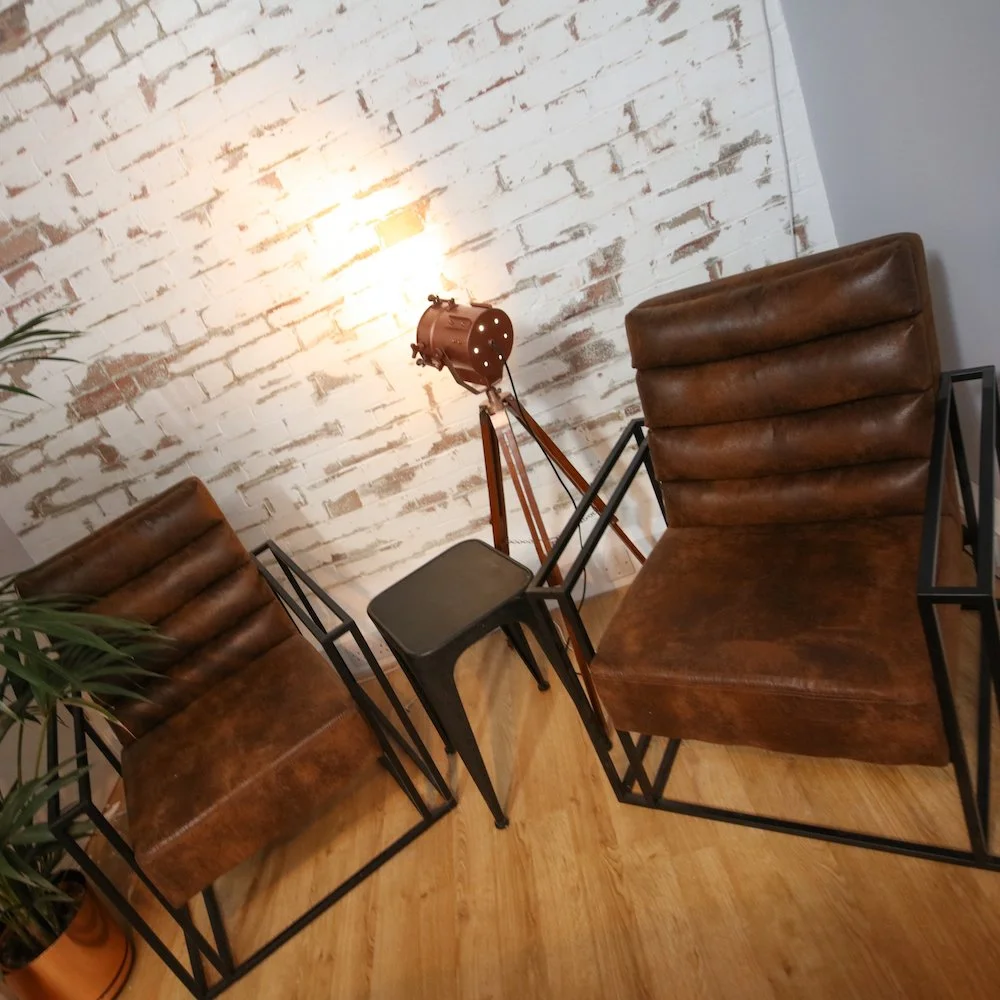 Two leather chairs with metal frames positioned on either side of a small black side table, with a vintage floor lamp shining light on a white brick wall in the background.