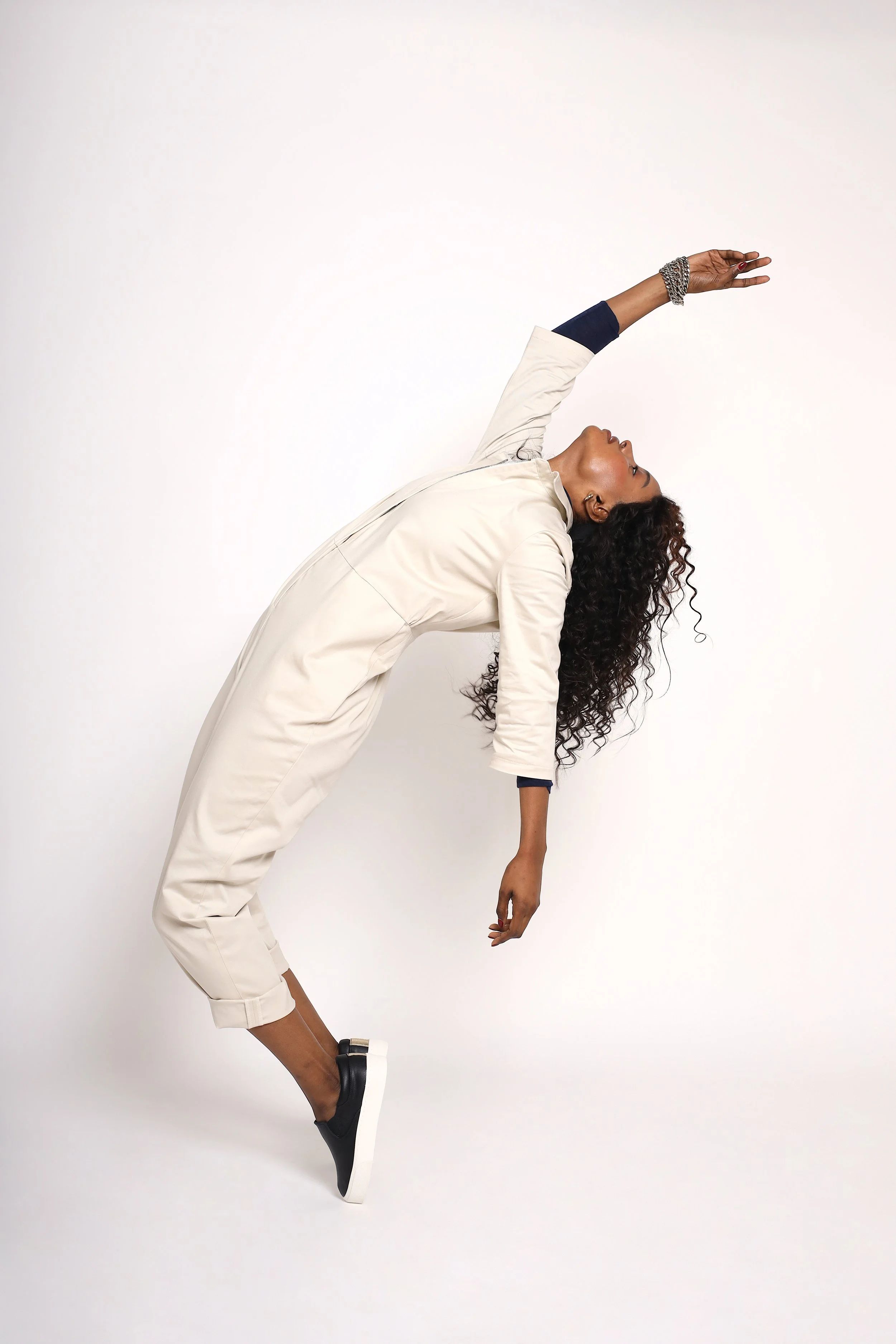 A woman with curly hair is in a pose where she is leaning backwards, with one arm reaching upward and the other hanging down, against a plain white background.