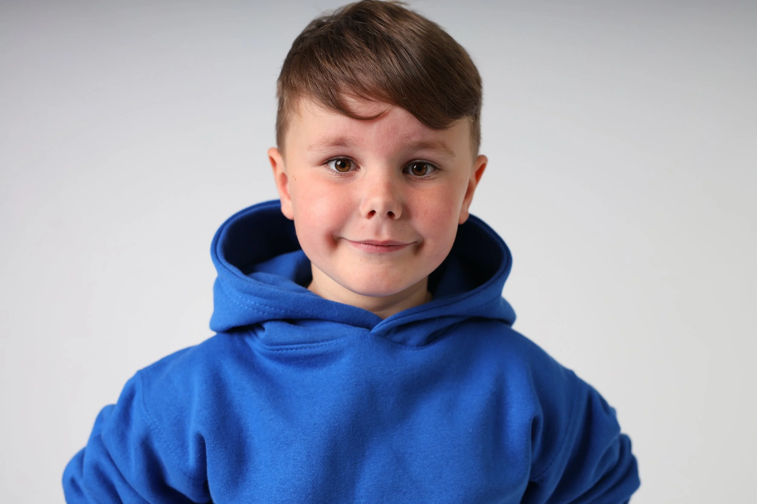A young boy with brown hair wearing a blue hoodie, smiling softly at the camera, against a plain white background.