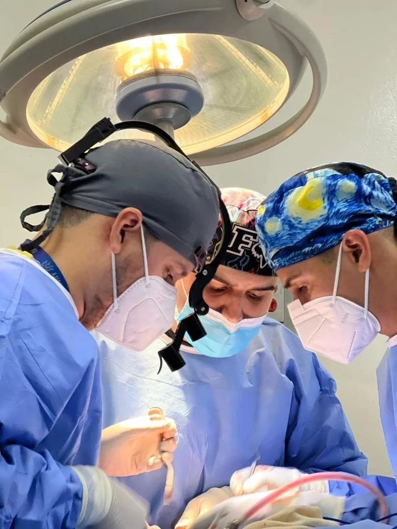 Three surgeons in scrubs and masks performing surgery under a surgical light.