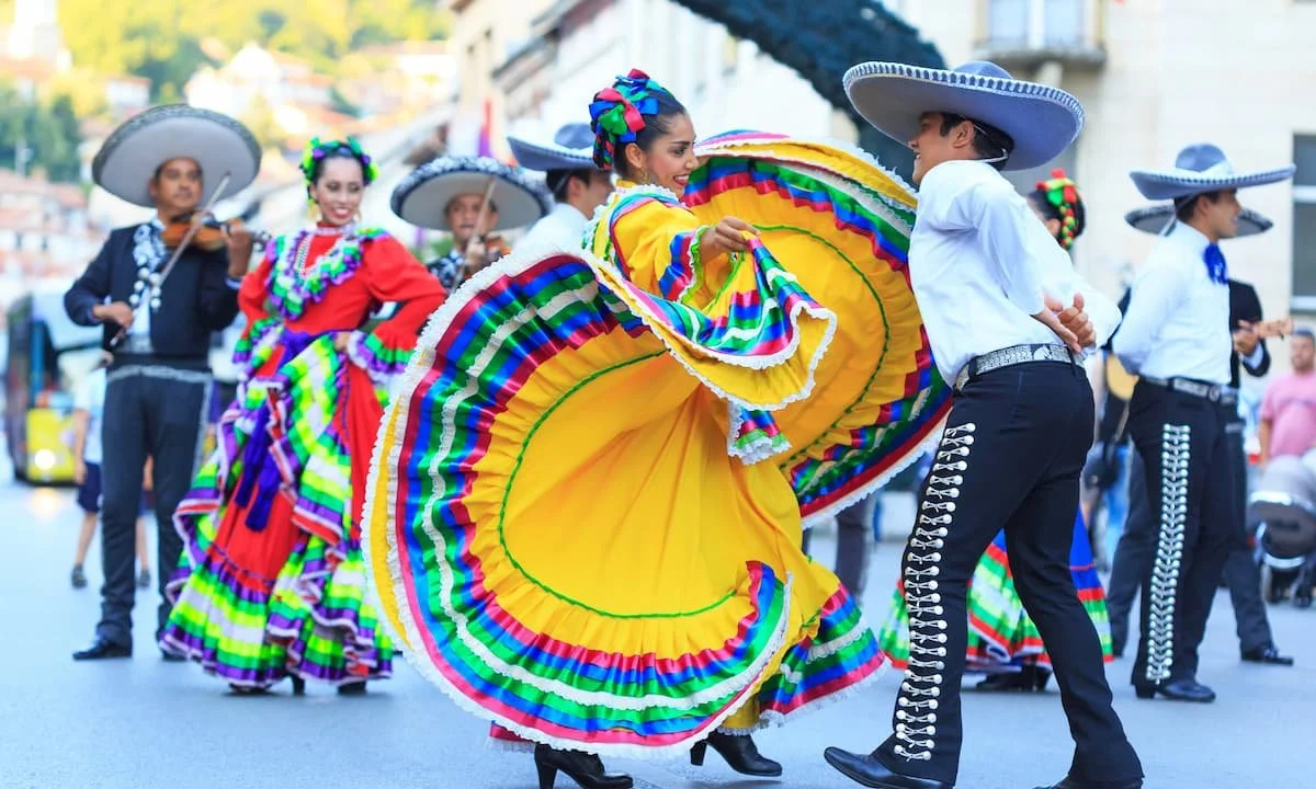 Mexican dancers in colorful traditional costumes perform during a celebration or parade in the street.