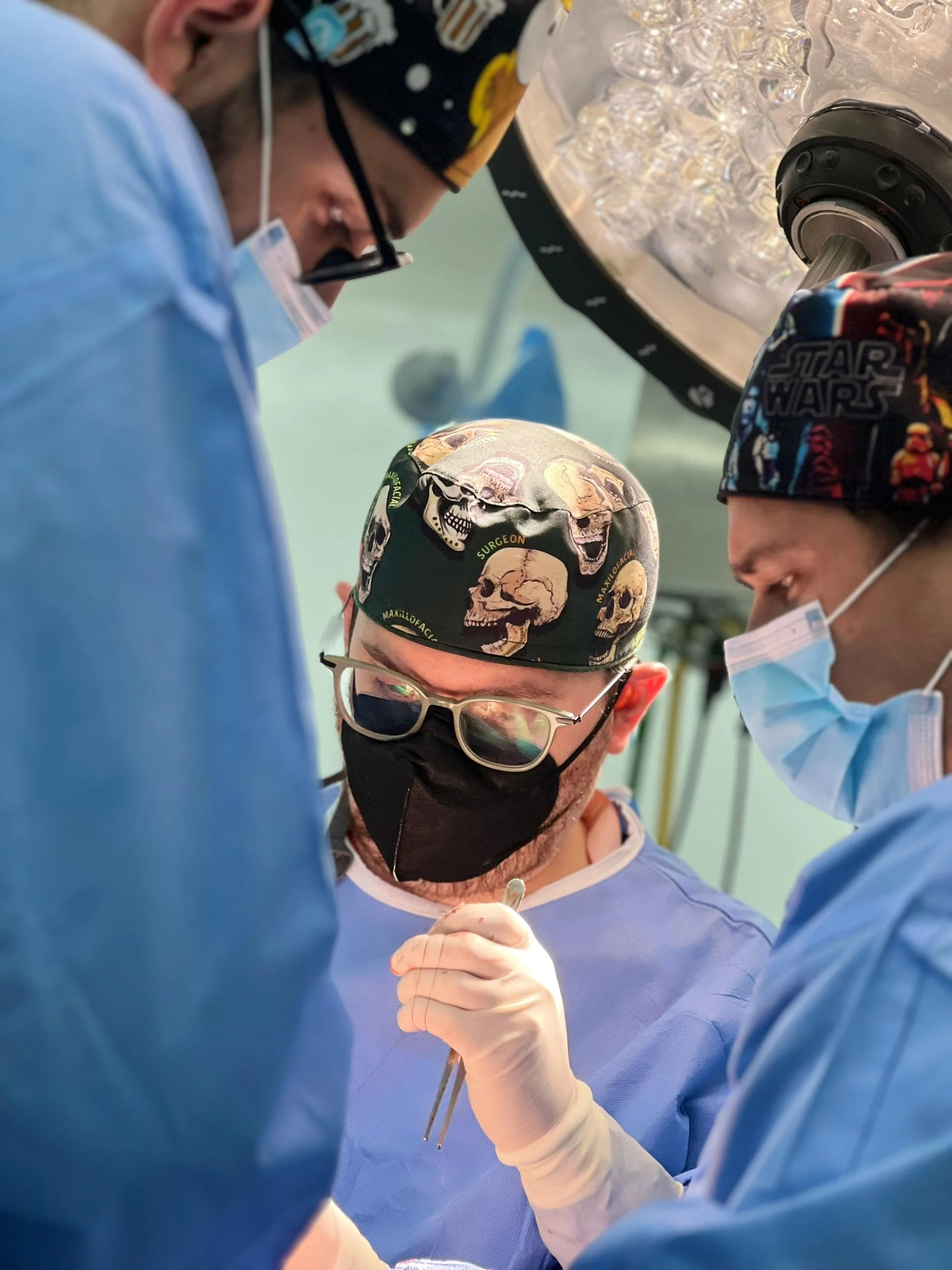 Surgeon in scrubs, wearing glasses, a skull-patterned surgical cap, and a black mask, performing surgery with assistants in blue scrubs, masks, and gloves under bright operating room lights.